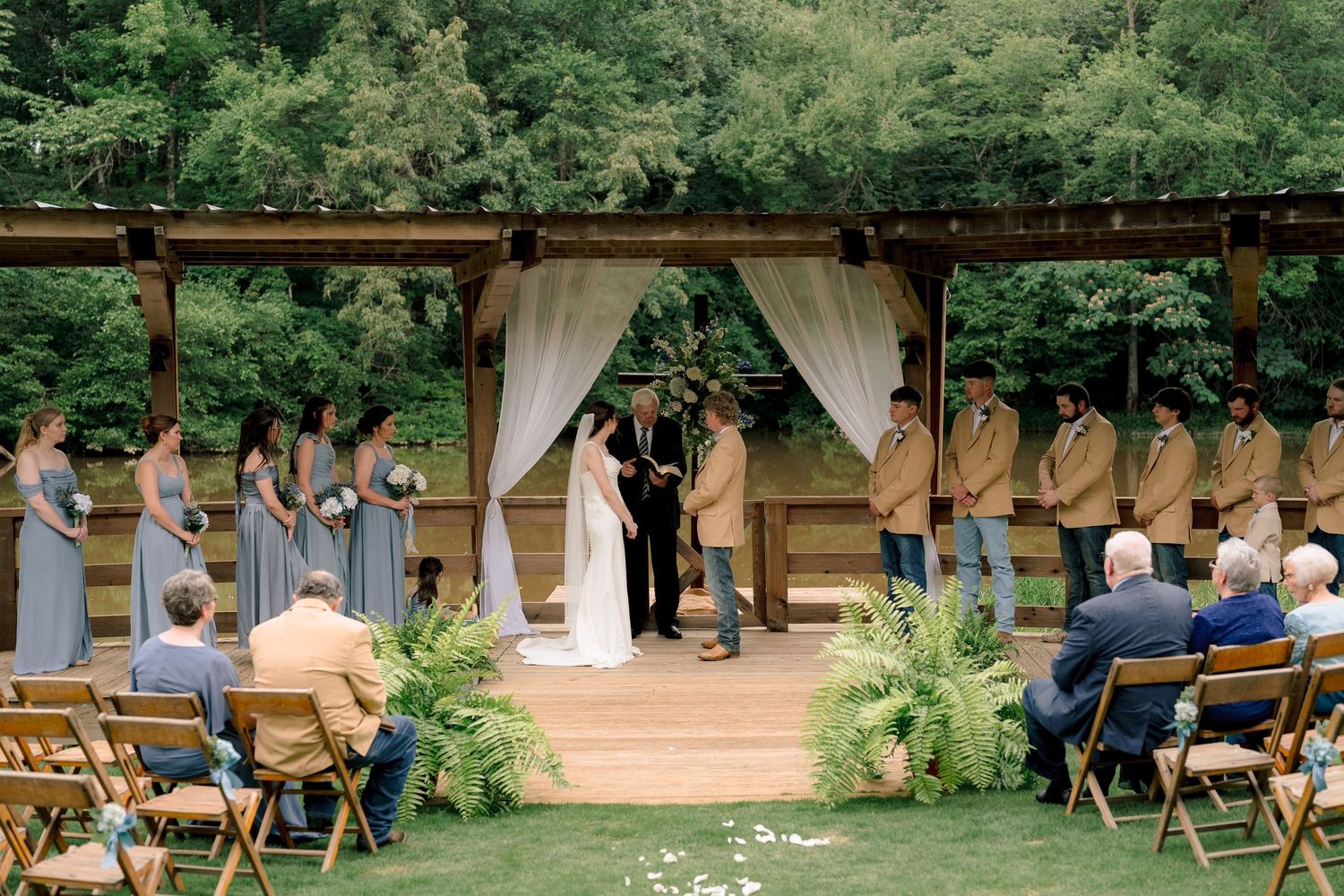 Elegant outdoor wedding ceremony at rustic wooden arch with full wedding party and seated guests on green lawn.