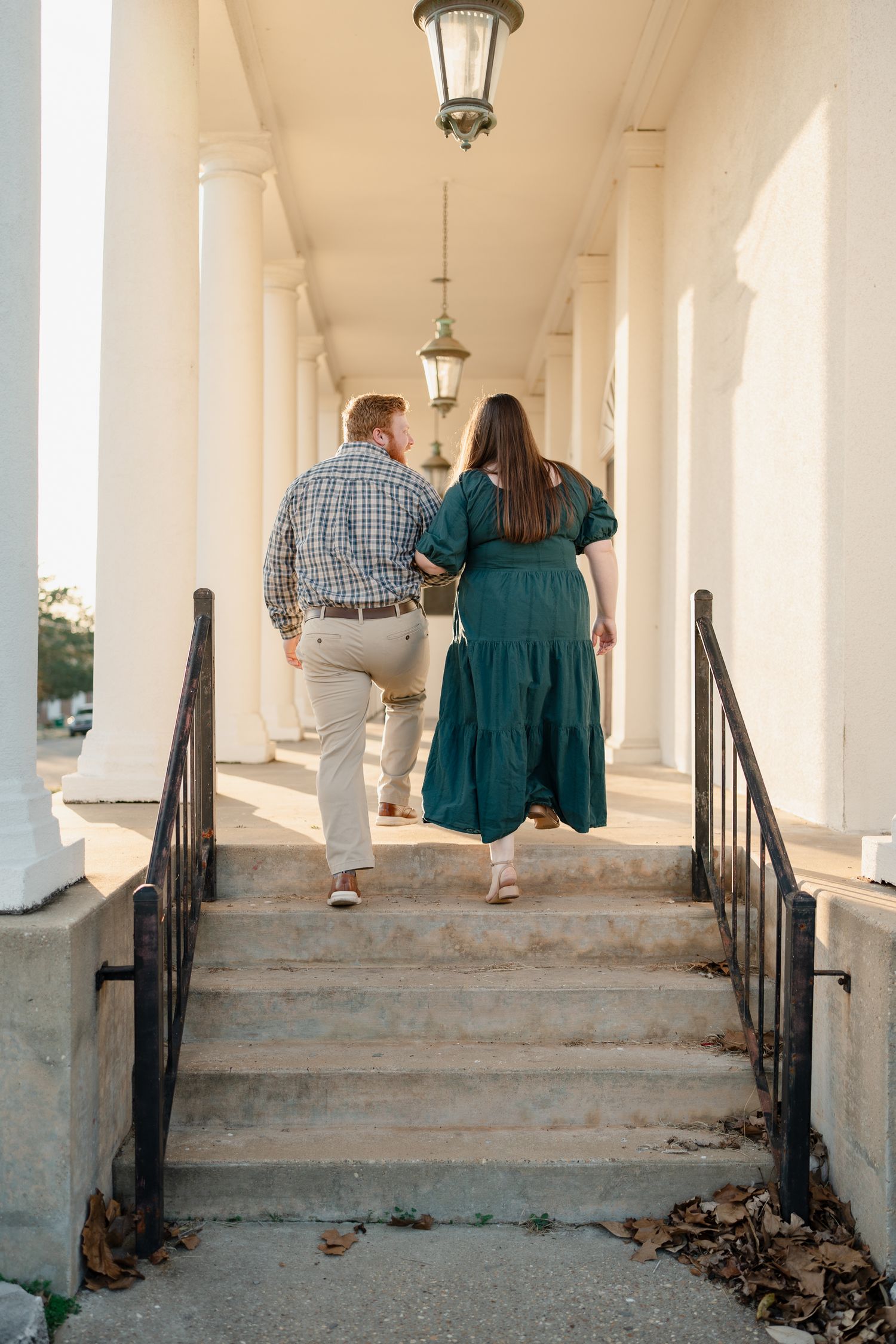 Couple walking up stone steps through a white columned corridor with hanging lanterns during golden hour.