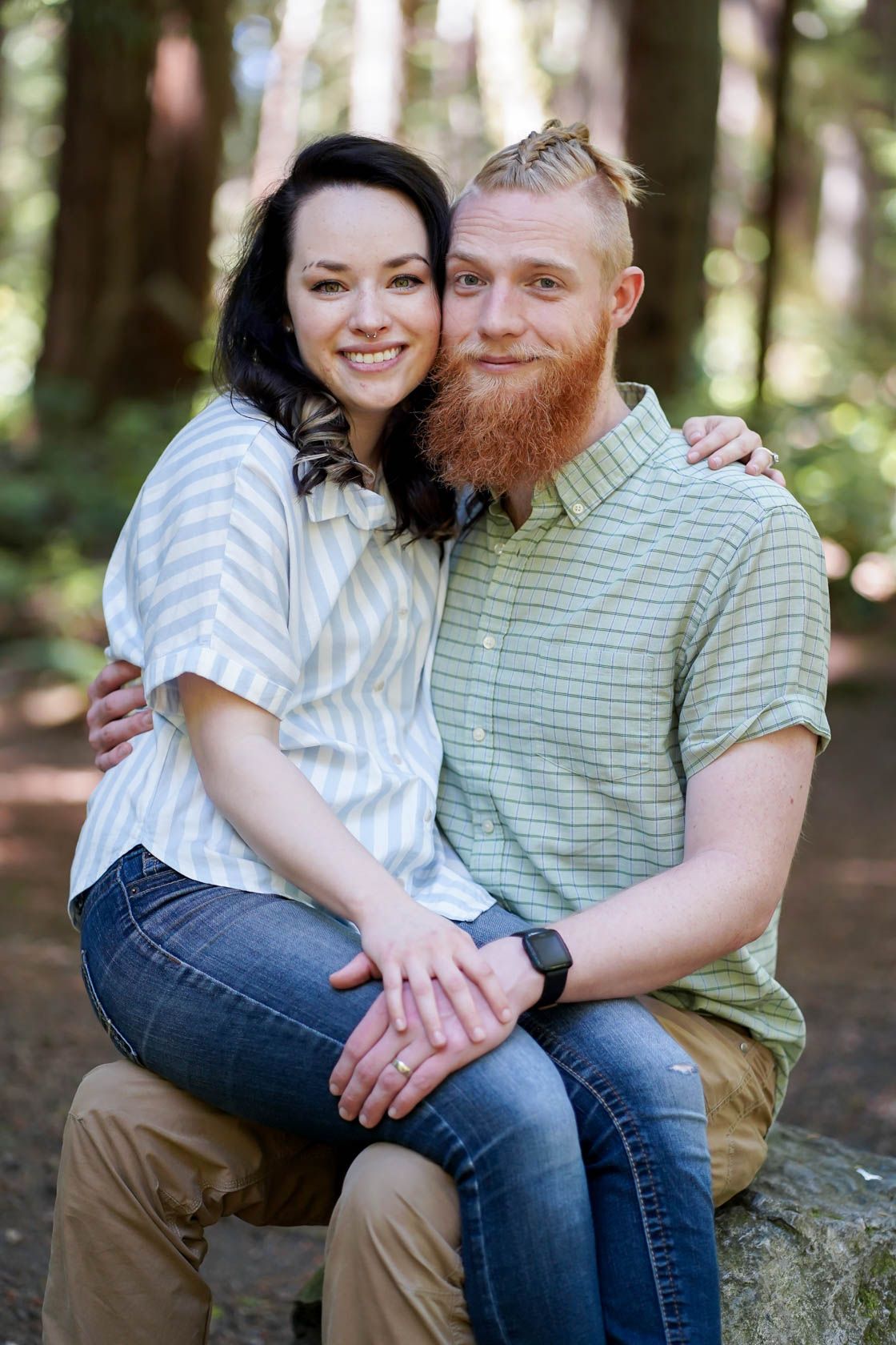 An intimate portrait of a couple in casual clothing sitting on a rock in the woods.