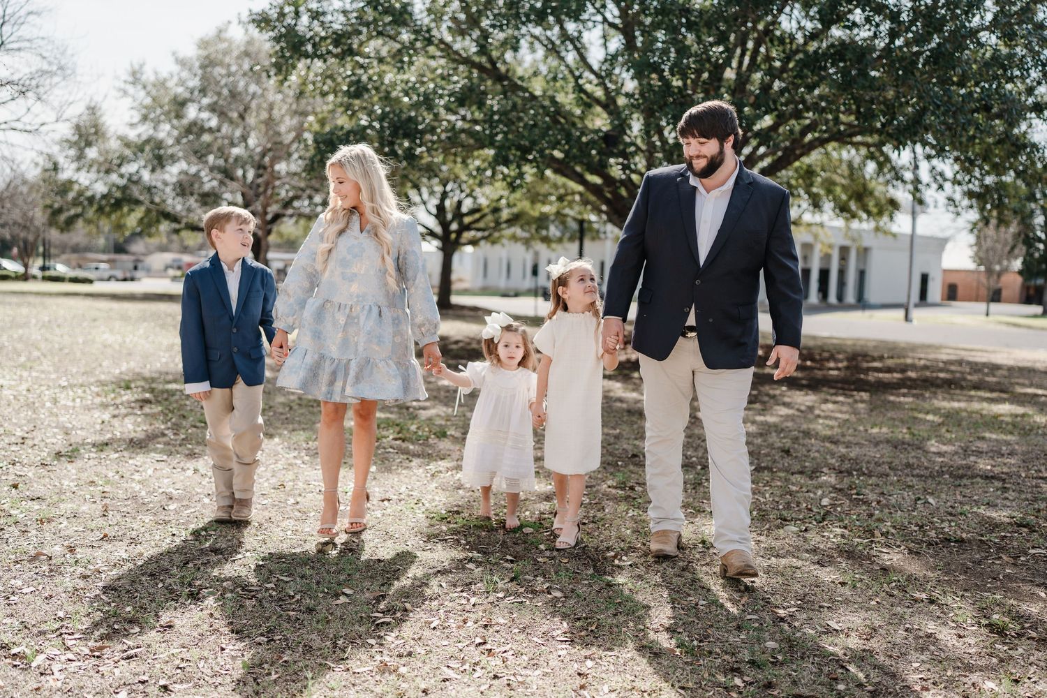 Family of five posed outdoors under large oak trees wearing coordinated light-colored outfits.