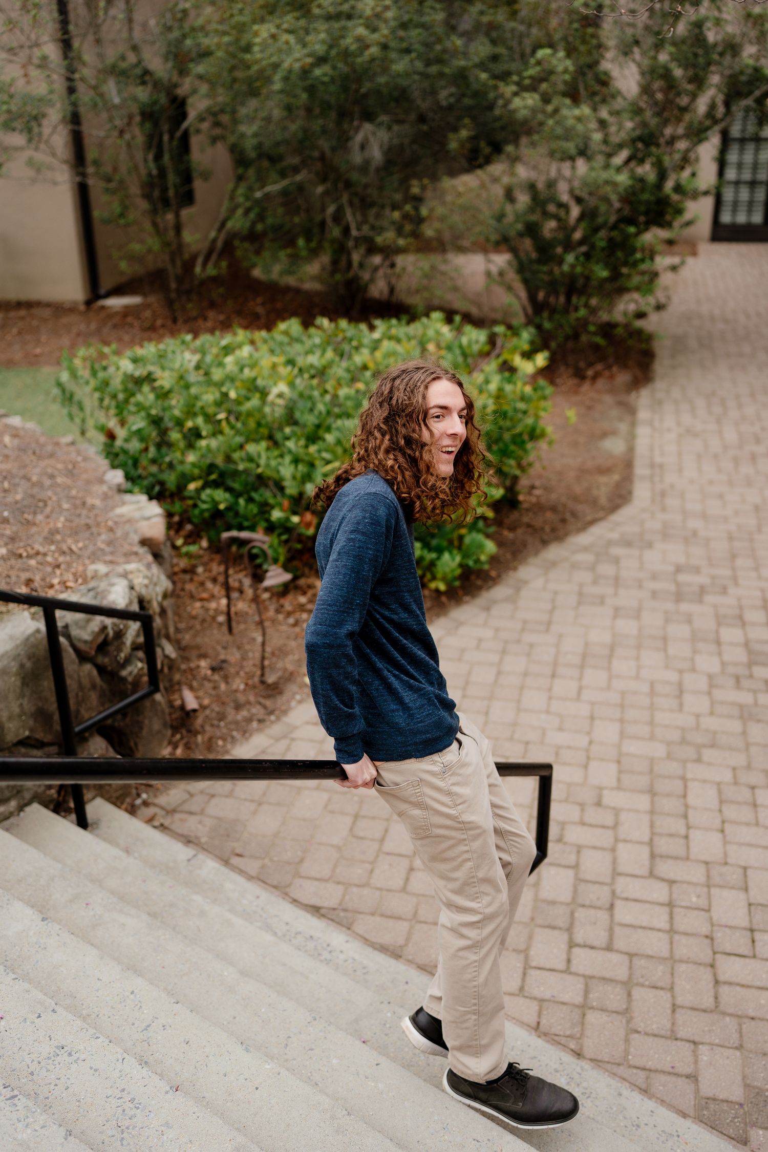 Person in navy sweater and khakis walking up brick steps near landscaped garden.