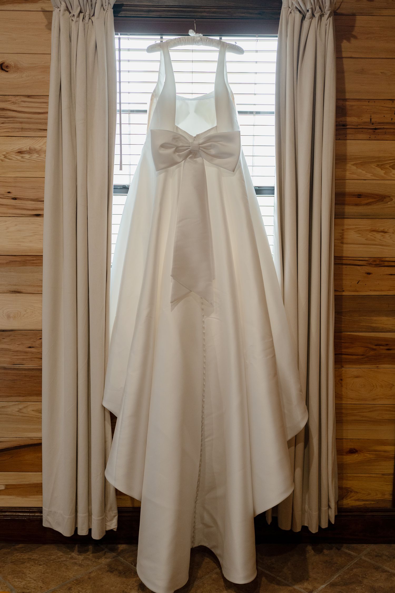 A white wedding dress hangs from a curtain rod against wooden paneled walls in a rustic setting.