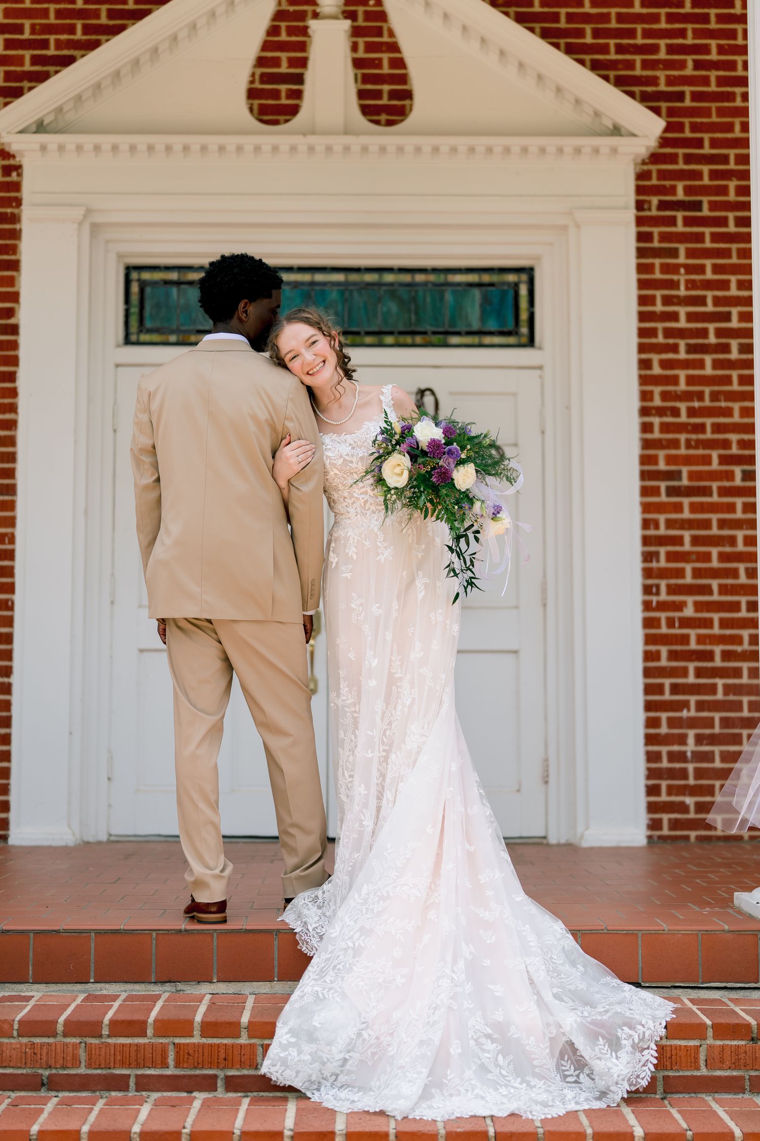 Smiling bride in lace gown embraces groom in tan suit on brick church steps holding floral bouquet.
