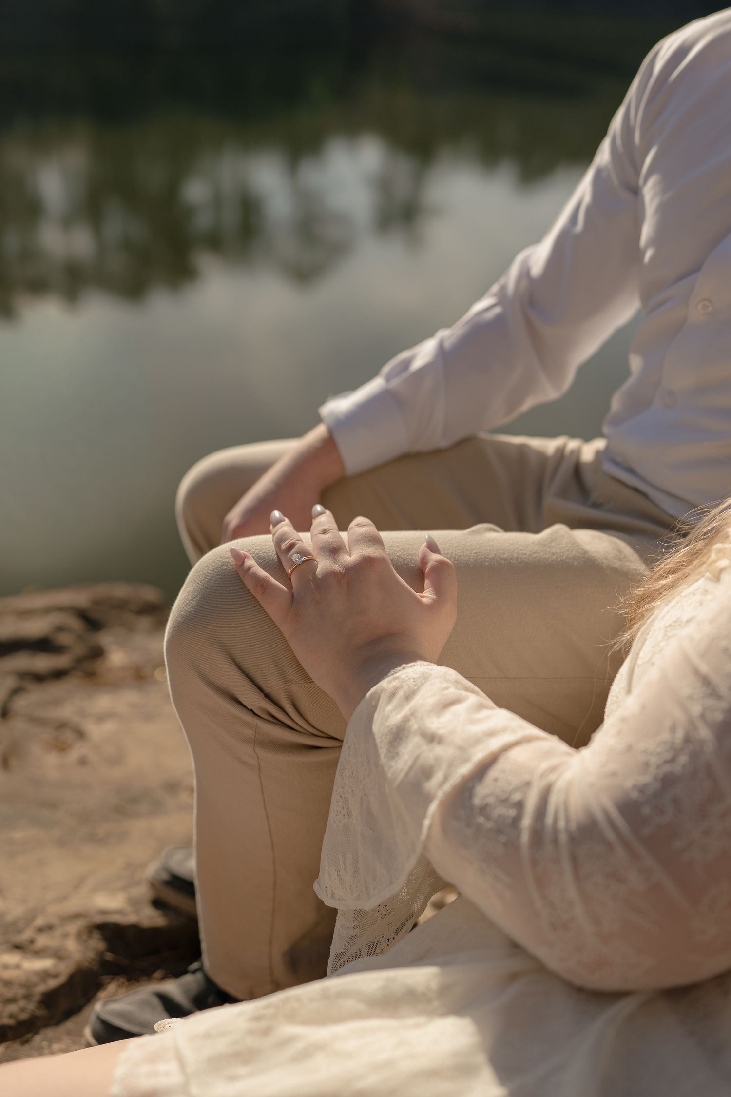 Close-up of a couple holding hands while sitting on rocks by a peaceful reflective lake outdoors.