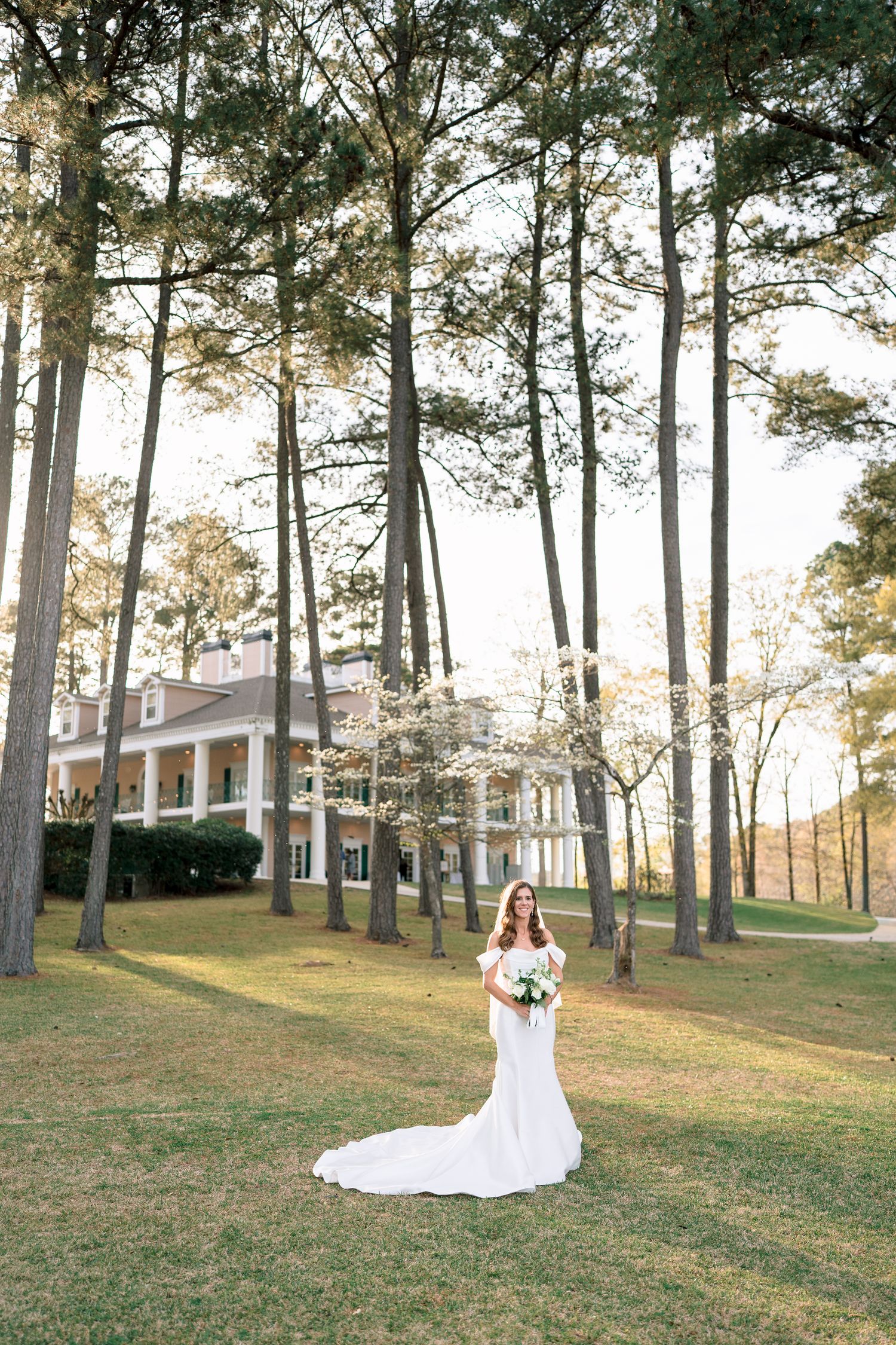 Bride in white mermaid gown holds bouquet among tall pines before a Southern mansion at golden hour.