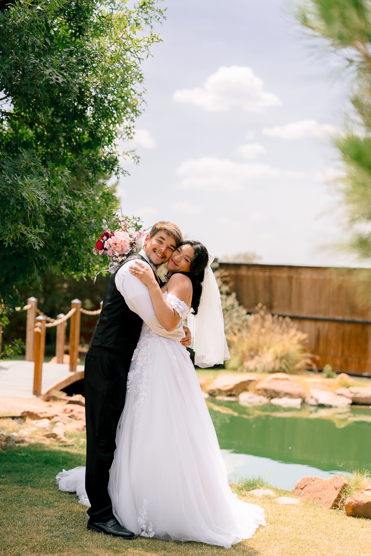 Happy newlyweds embrace near a garden pond, bride in white gown holding a bouquet while groom smiles joyfully.