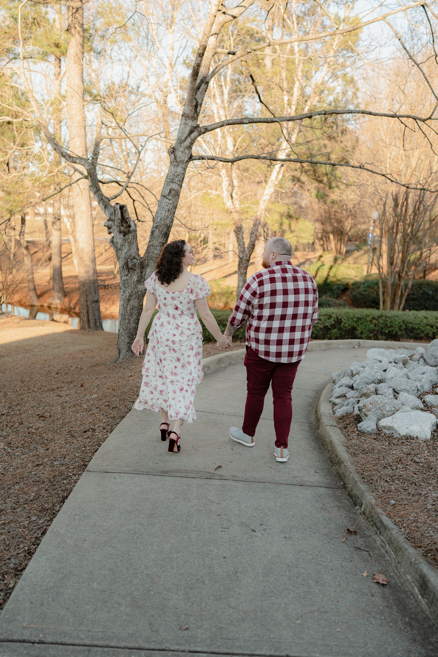 Two people walking together on a paved path through a park during autumn with fallen leaves on the ground.