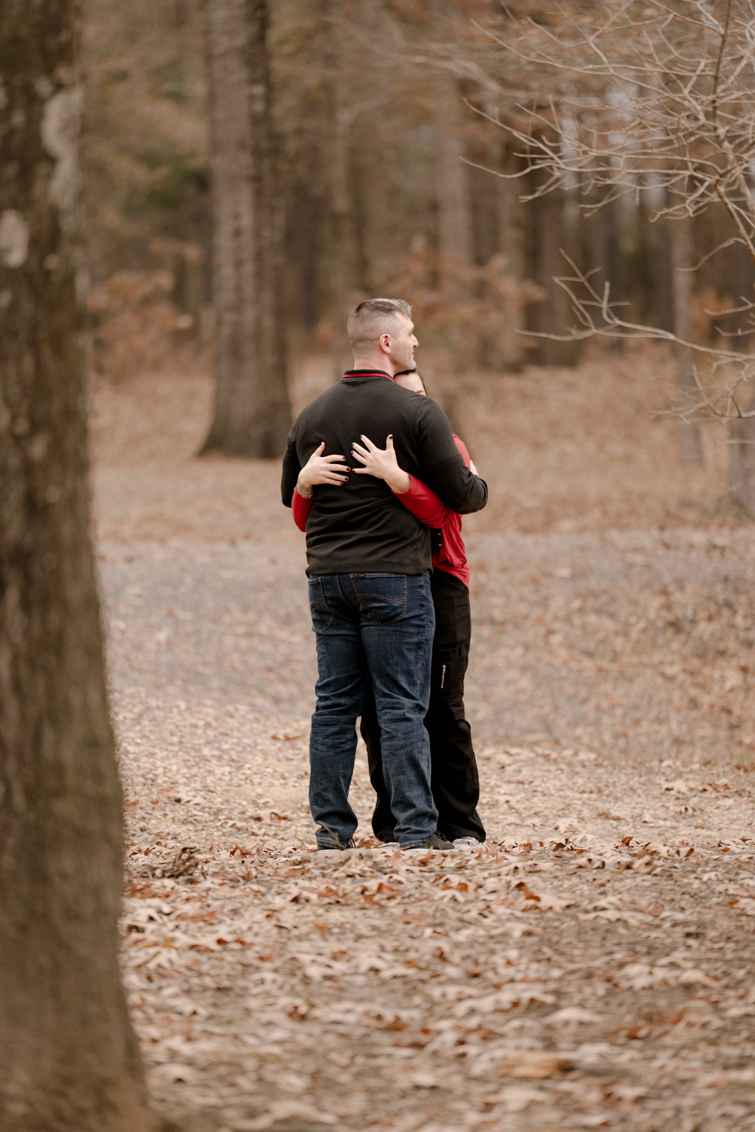 A couple embraces while standing on a leaf-covered path in an autumn forest with bare trees.