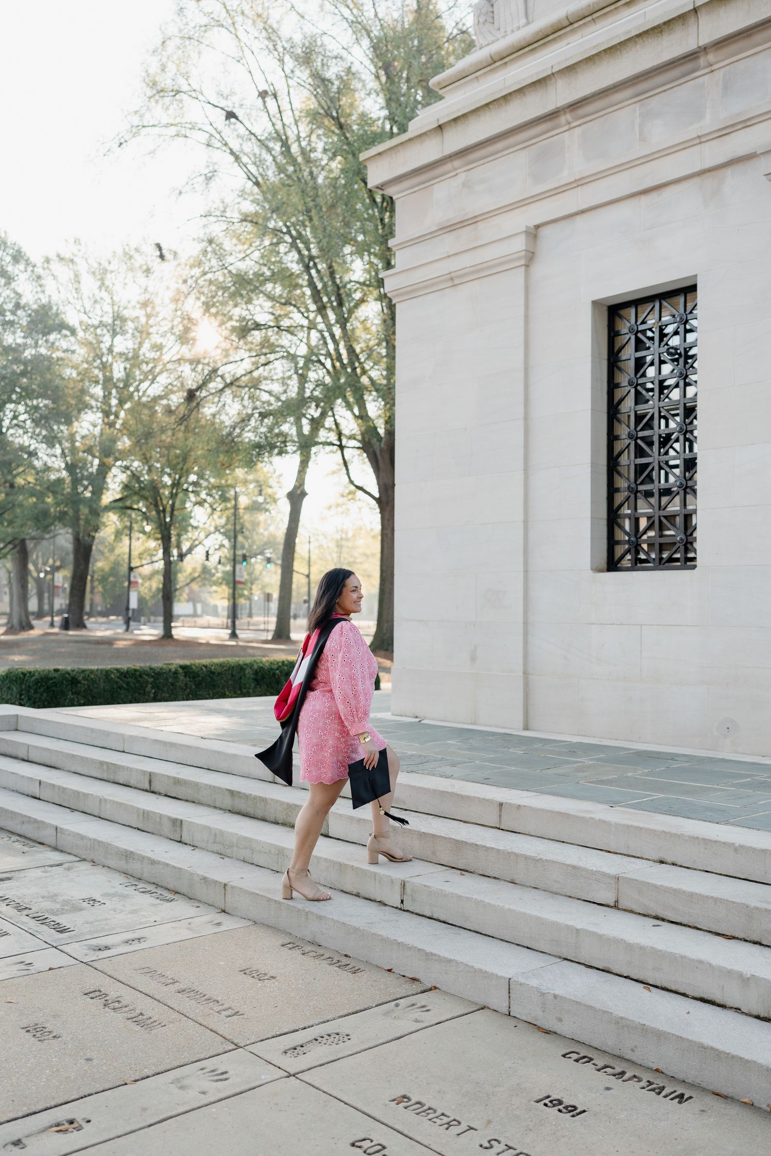 Graduate ascending white stone steps in pink dress with architectural details visible.