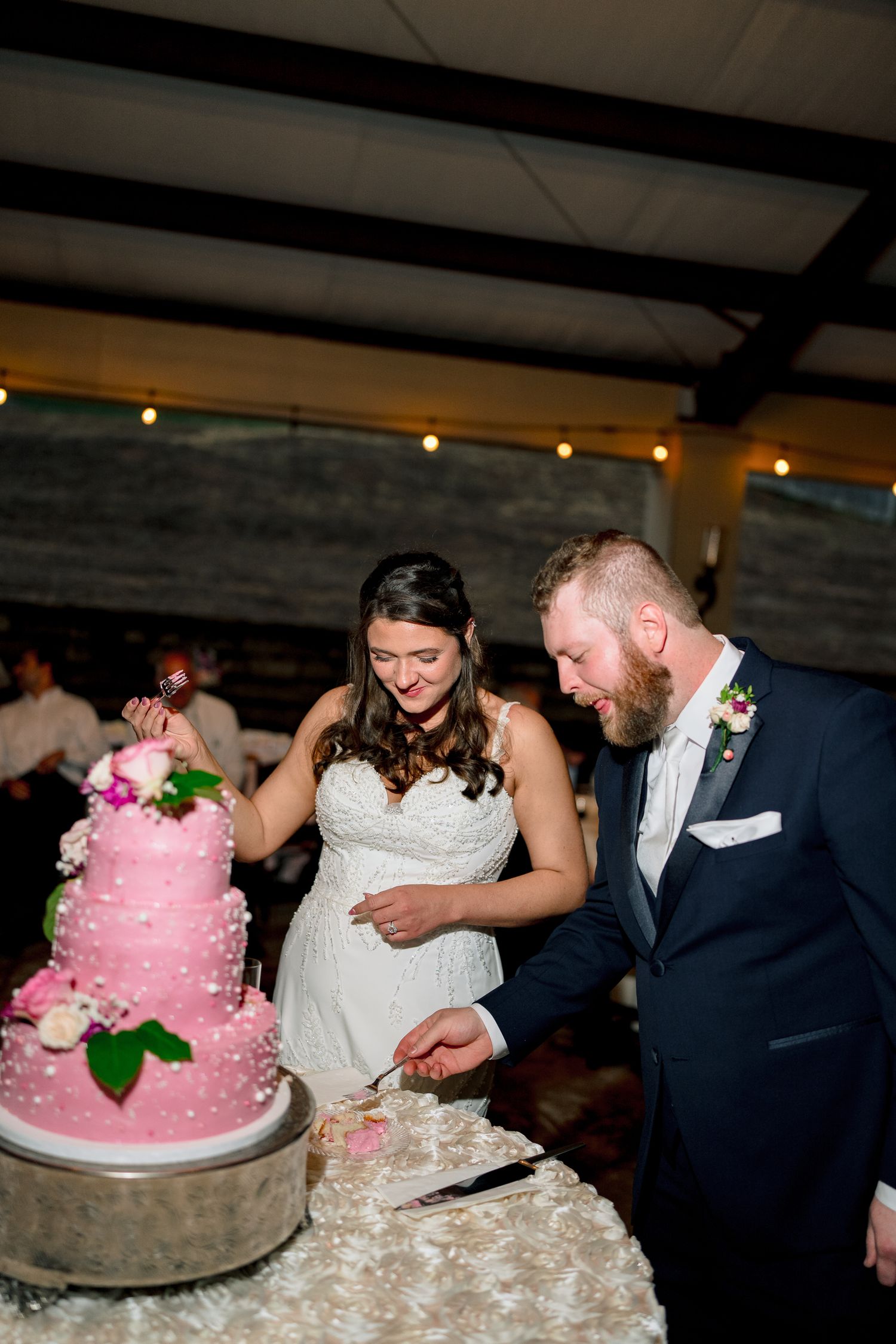 Bride and groom cut their three-tier pink pearl wedding cake together during indoor reception under string lights.