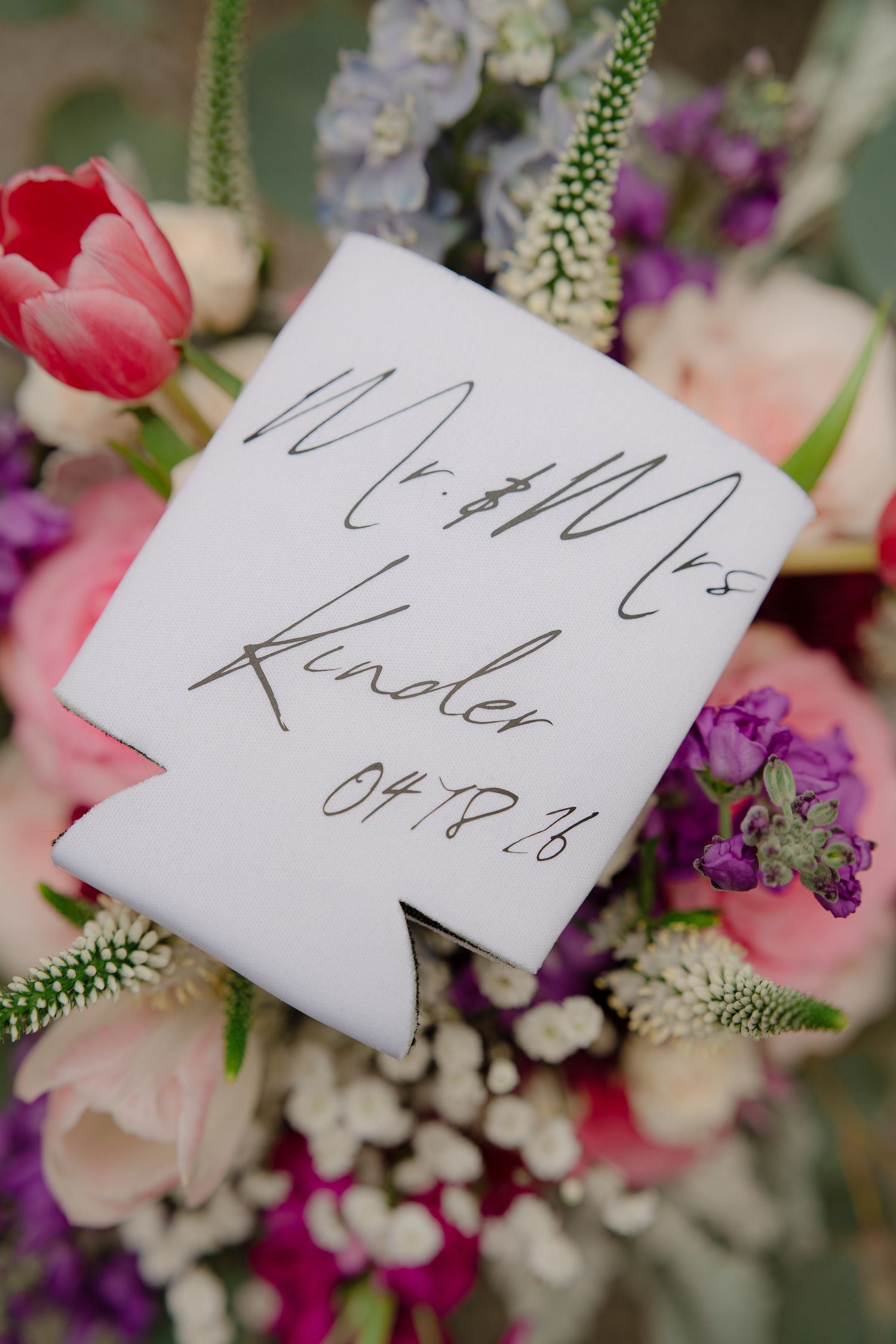 White wedding koozie for Mr. & Mrs. Kinder dated 04.18.16 resting on a colorful bridal bouquet.