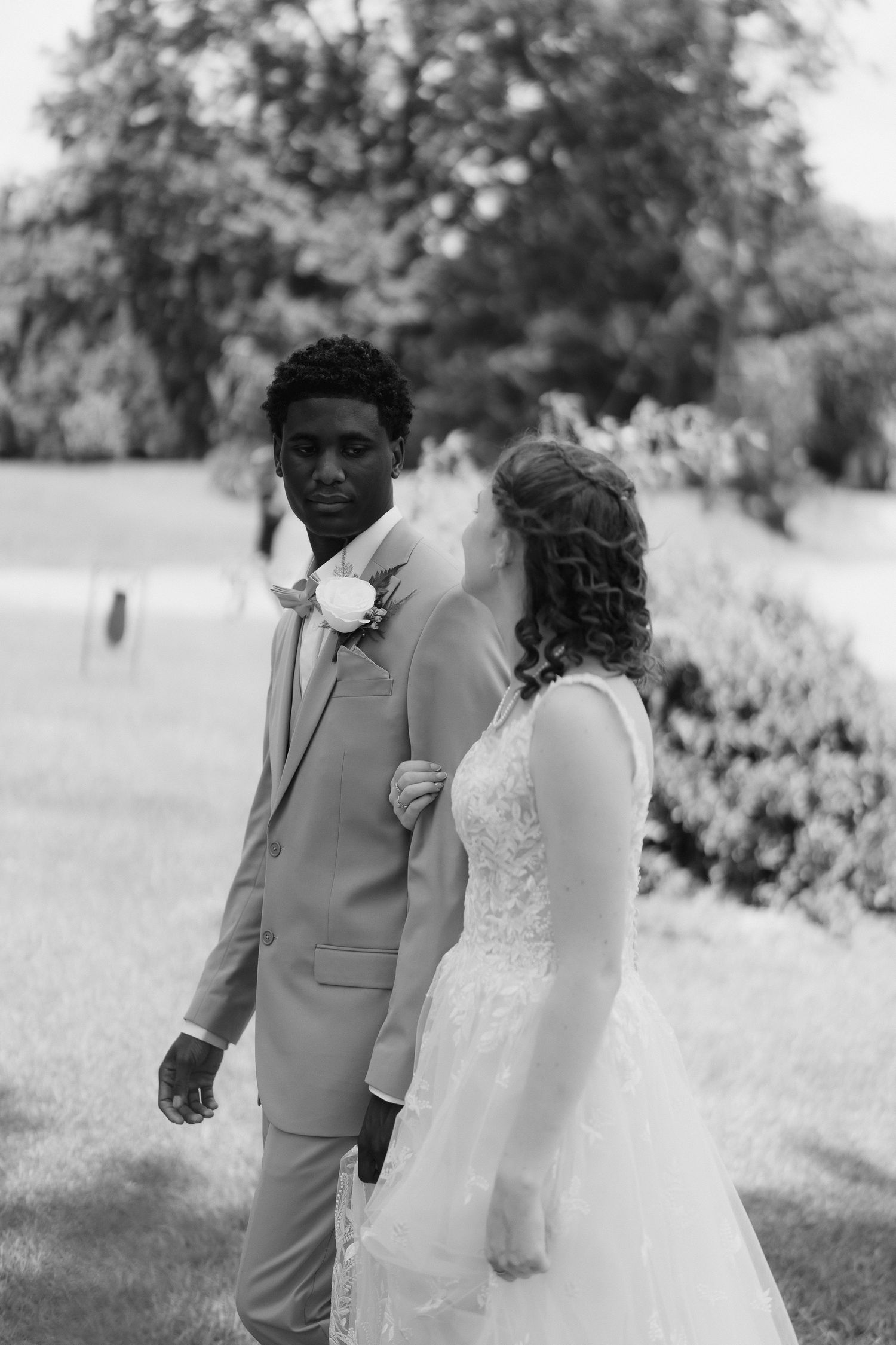 Black and white photo of interracial wedding couple embracing outdoors, groom in tan suit, bride in lace gown.