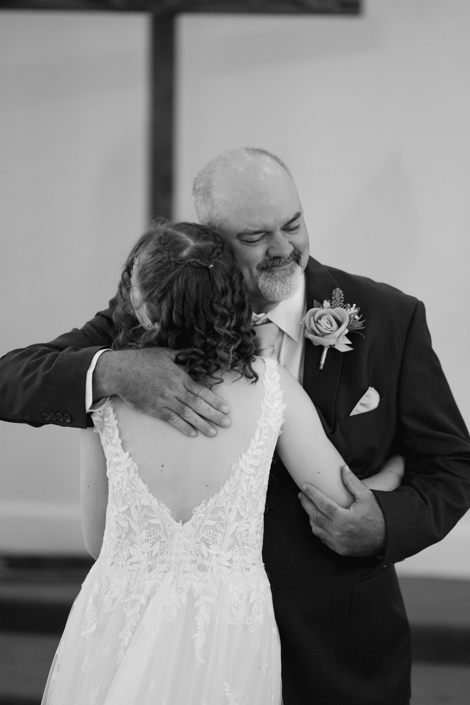 Black and white photo of bride in lace backless gown embracing older man in dark suit with floral boutonniere at wedding.