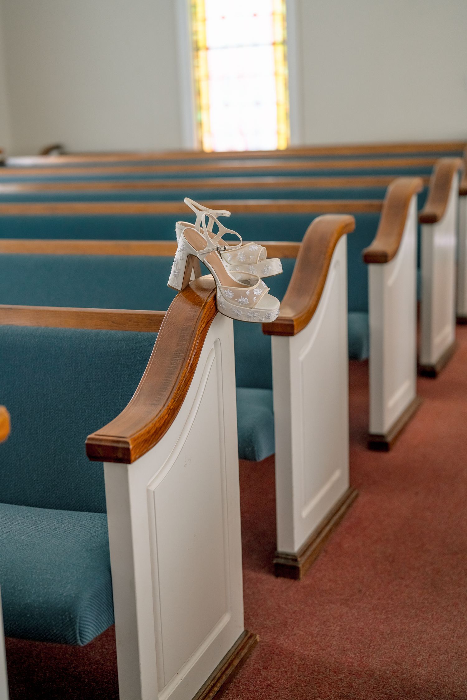 White bridal heels displayed on wooden church pew with teal cushions before wedding ceremony.