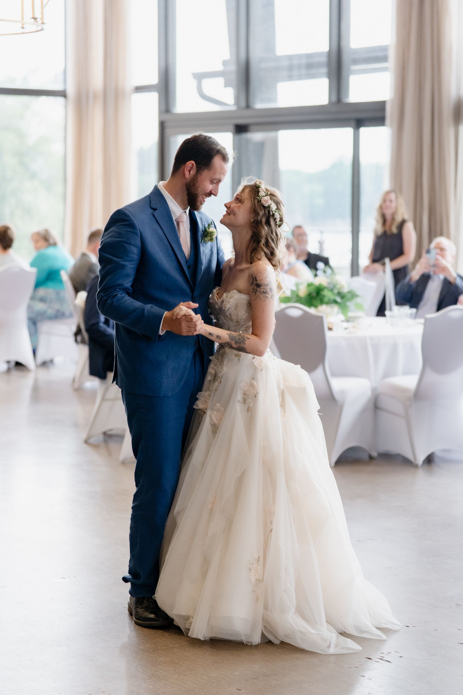 Couple dancing together in a bright reception hall with white-covered chairs and natural lighting.