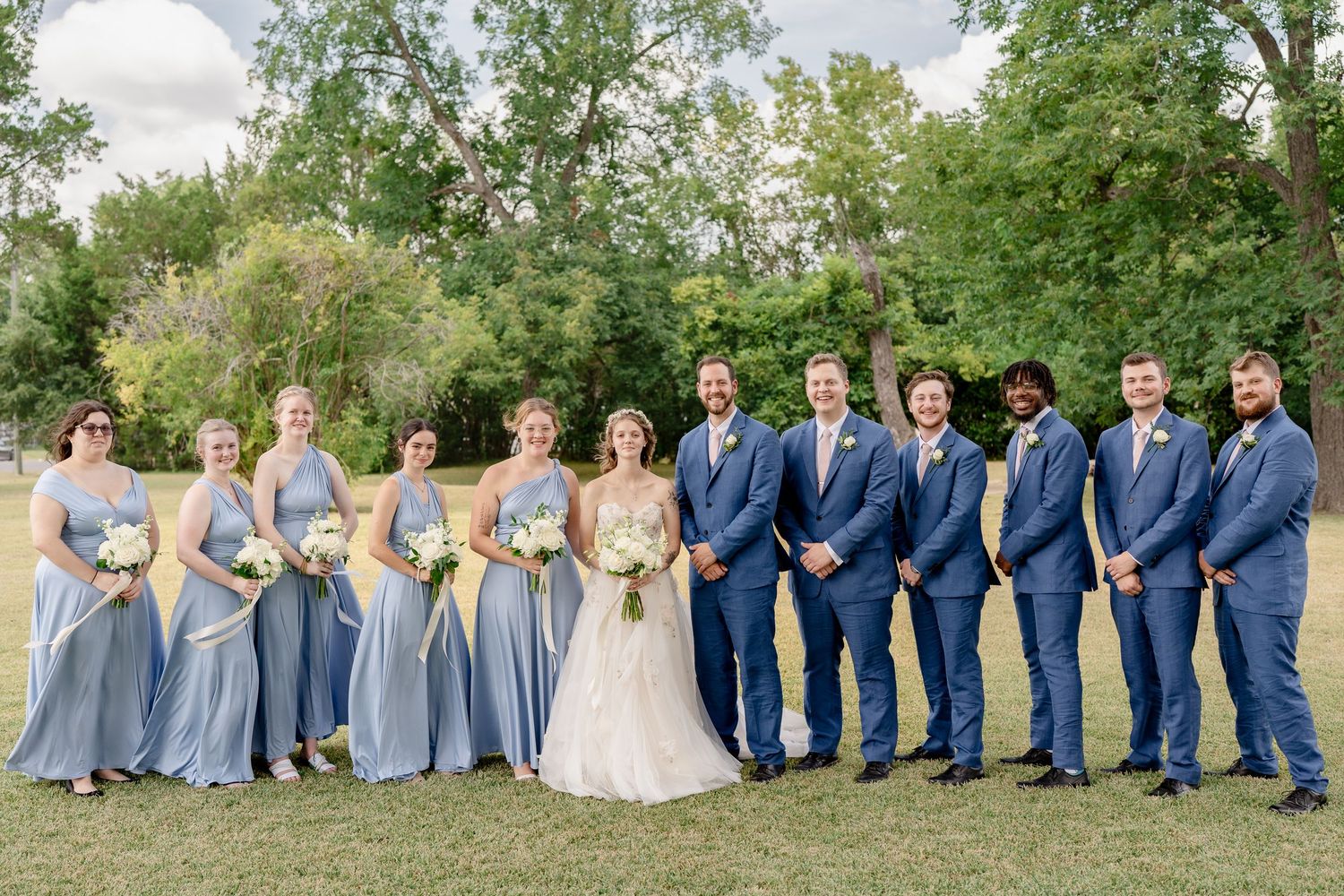 Wedding party in blue and gray attire poses together on grassy field during outdoor celebration.