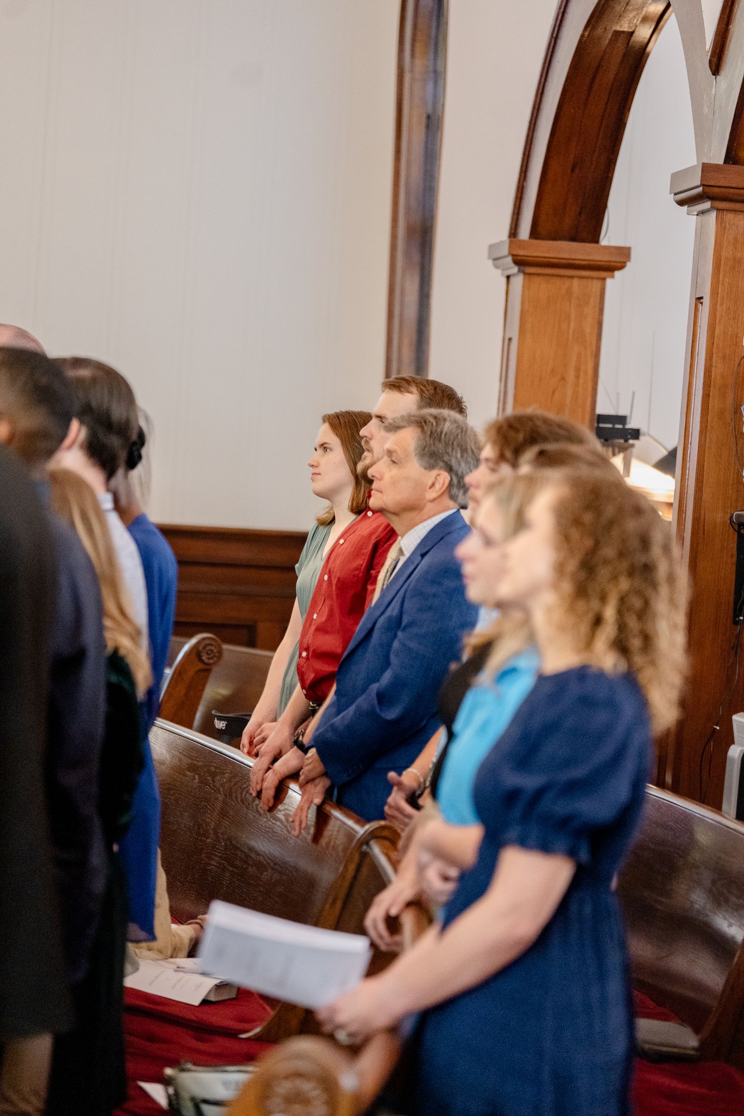 A group of wedding guests stand in church pews during a ceremony, wearing formal attire in shades of blue.