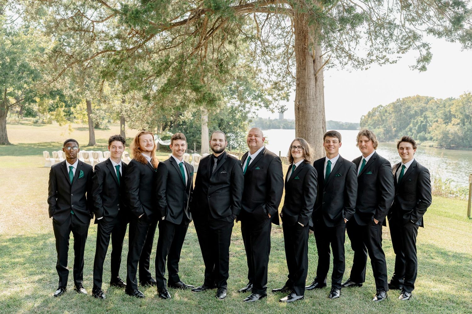 Wedding party lined up in formal black suits and emerald dresses against a natural outdoor backdrop.