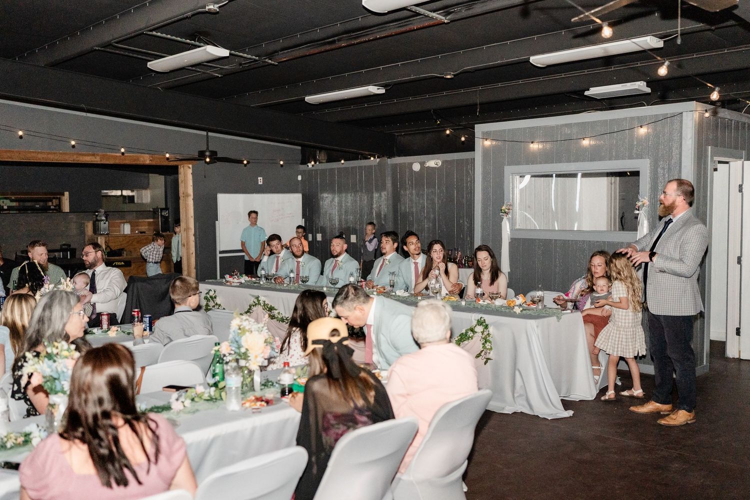 A wedding reception in a modern venue with guests seated at decorated tables while a speaker addresses the room.