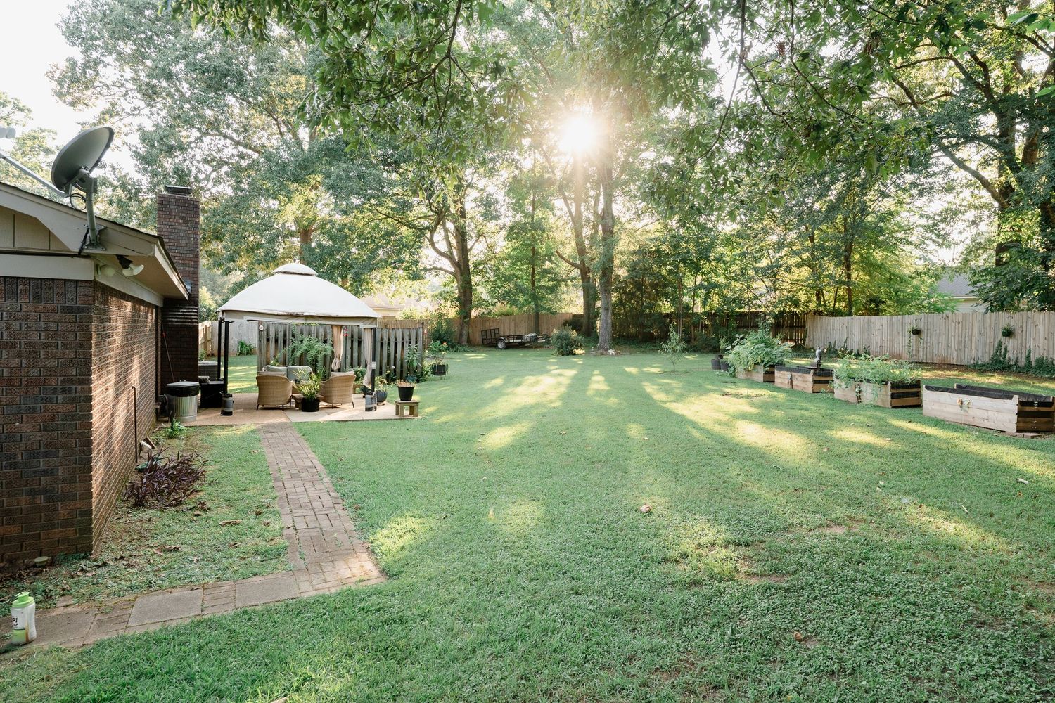 Sunlit backyard garden with brick pathway leading to white gazebo and lush green lawn surrounded by trees.
