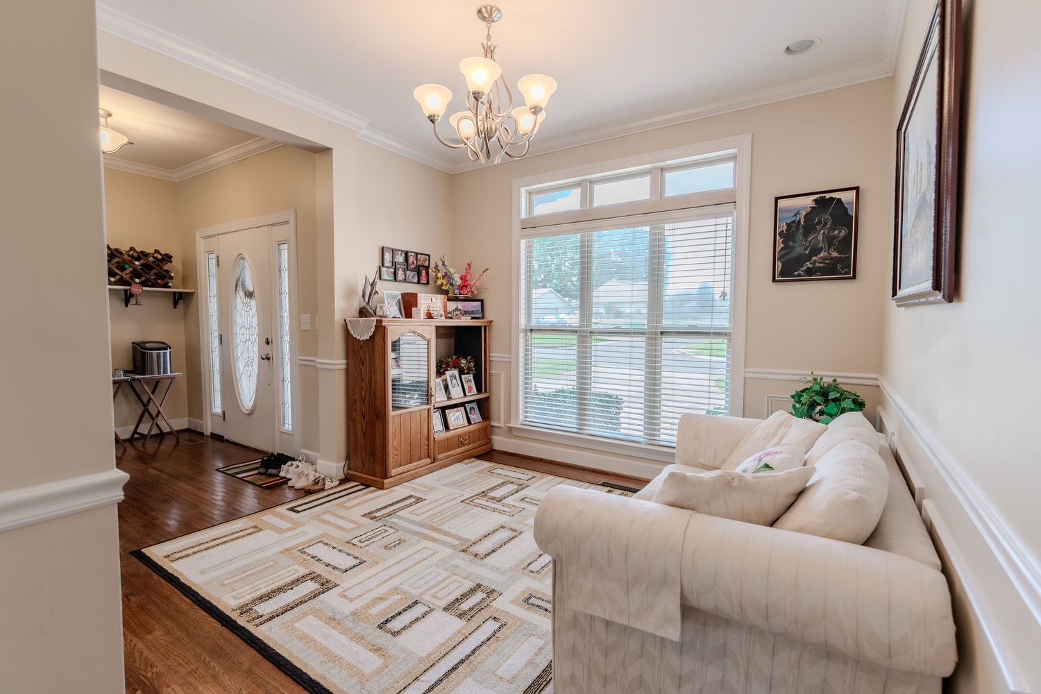 A bright modern living room features white walls, hardwood floors, and a crystal chandelier above plush seating.