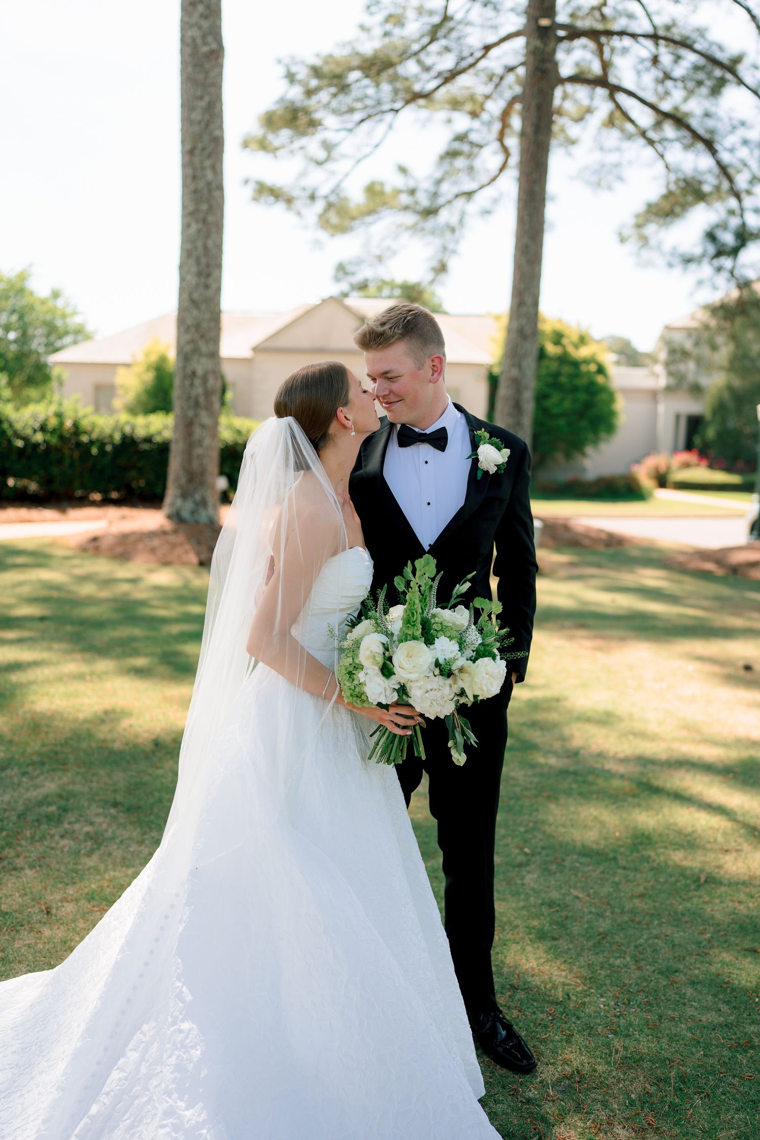 Bride in white gown with veil and groom in black tuxedo share a romantic moment outdoors holding white floral bouquet.