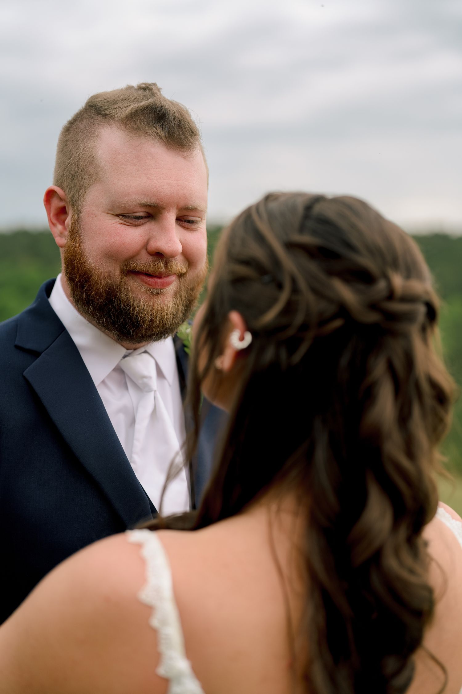 Groom in navy suit smiling tenderly at bride during outdoor wedding ceremony with green hills in background.