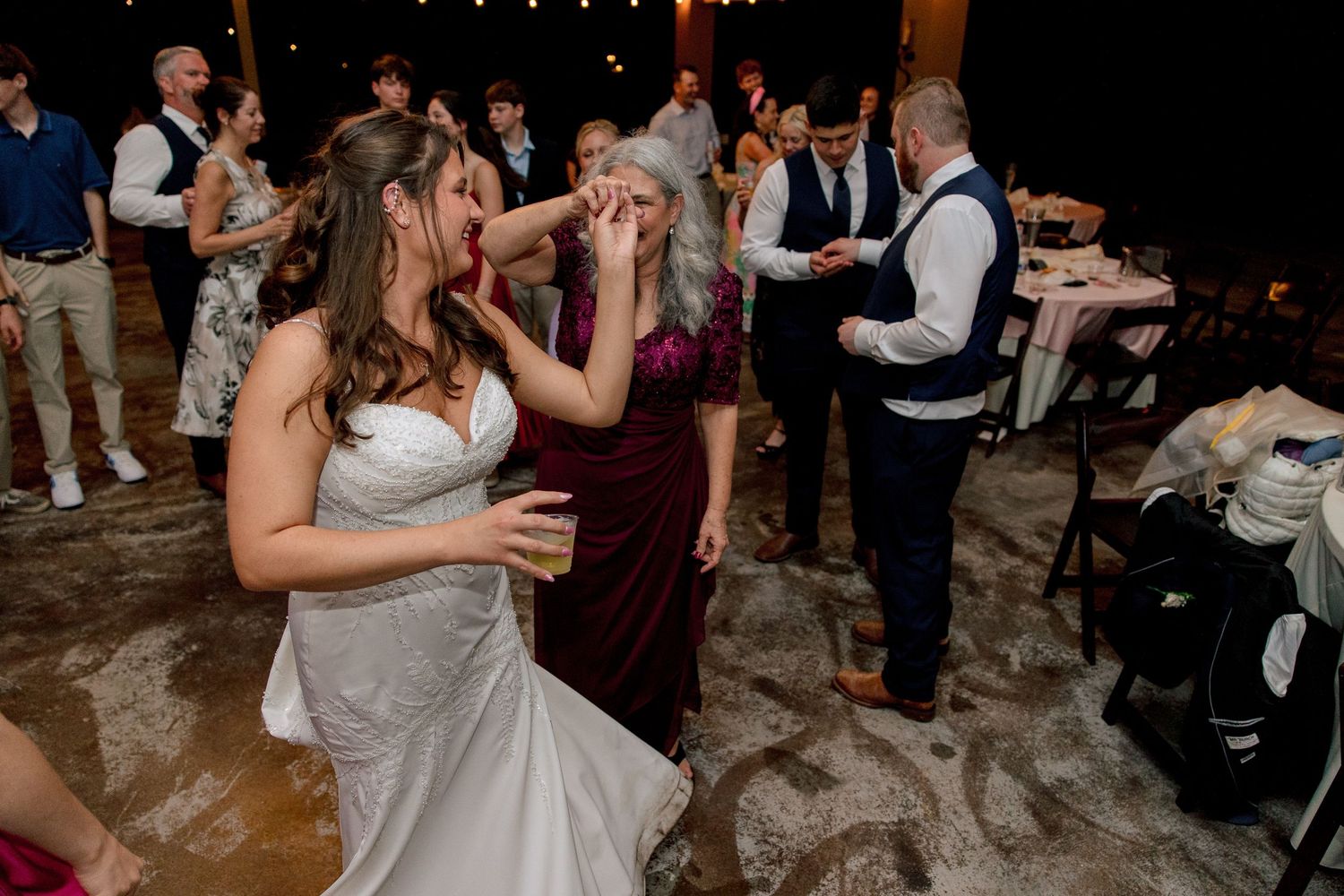 Bride in white gown laughs and dances with a guest in burgundy dress during lively wedding reception.