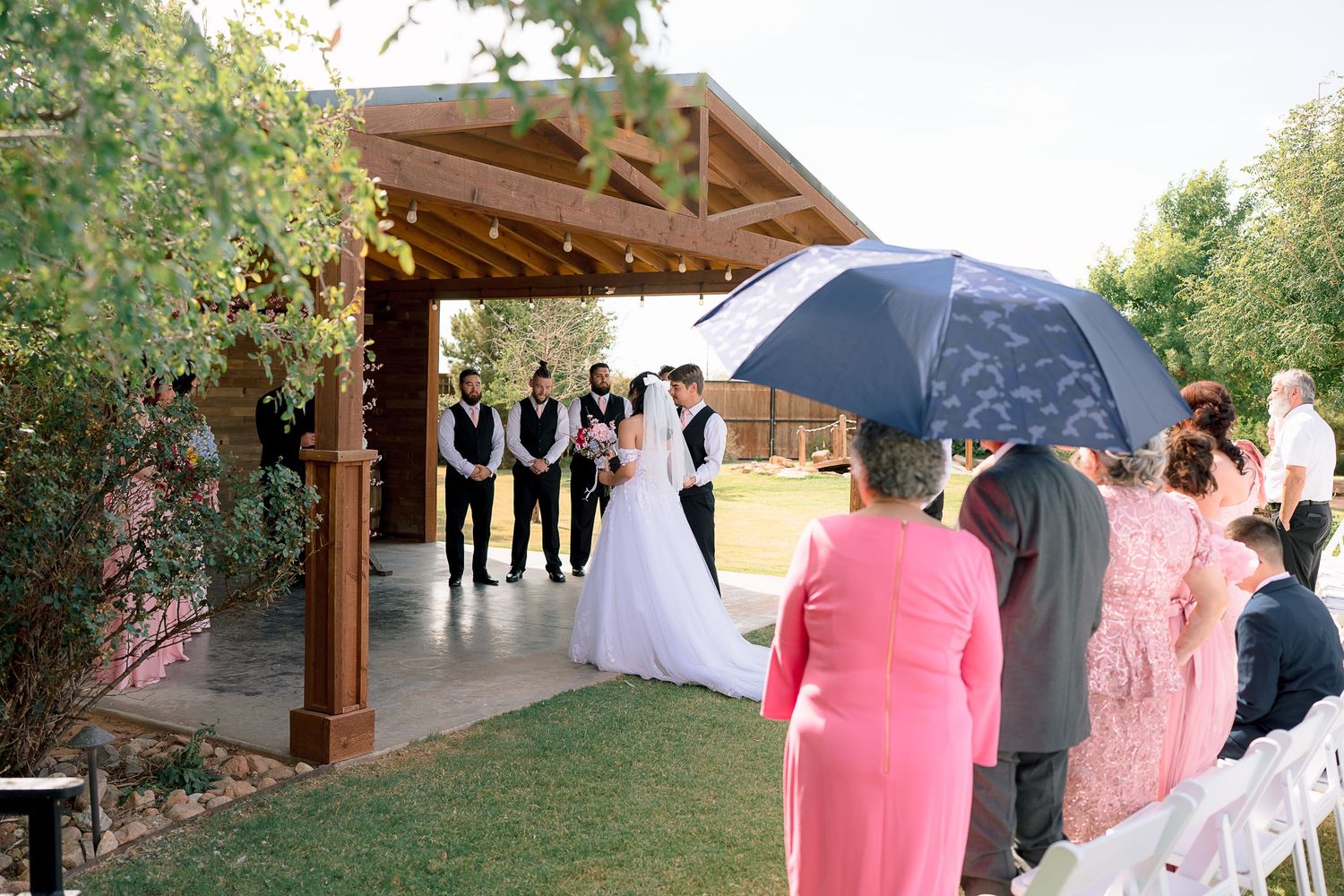 Outdoor wedding ceremony under wooden pergola with bride, groom, and groomsmen, guests seated on white chairs.