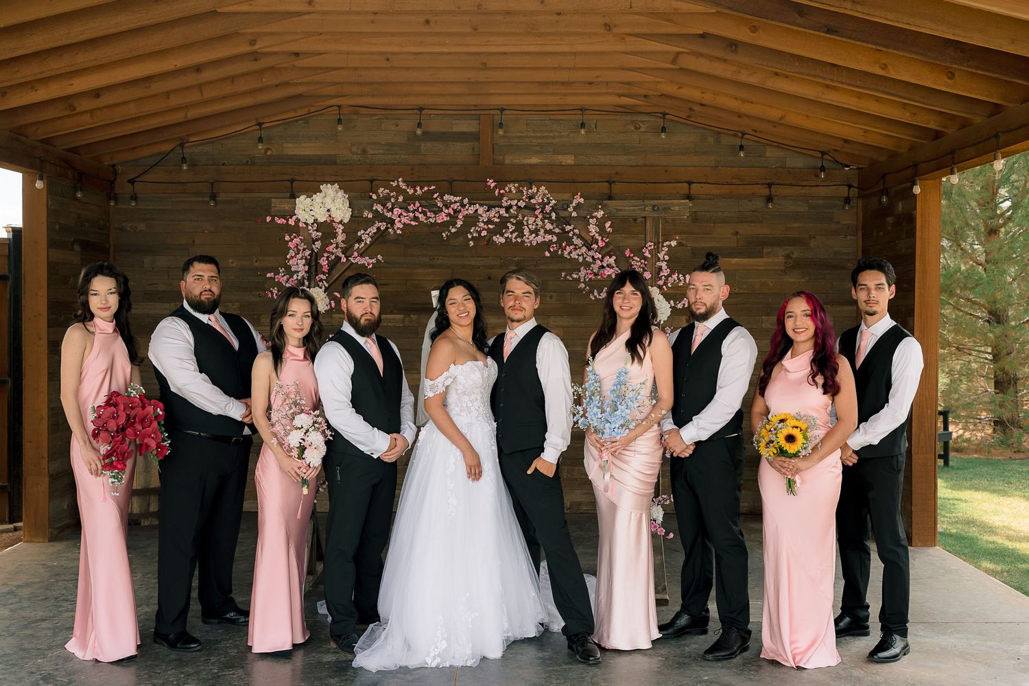 Wedding party posing under floral arch at rustic barn venue in pink and black attire.