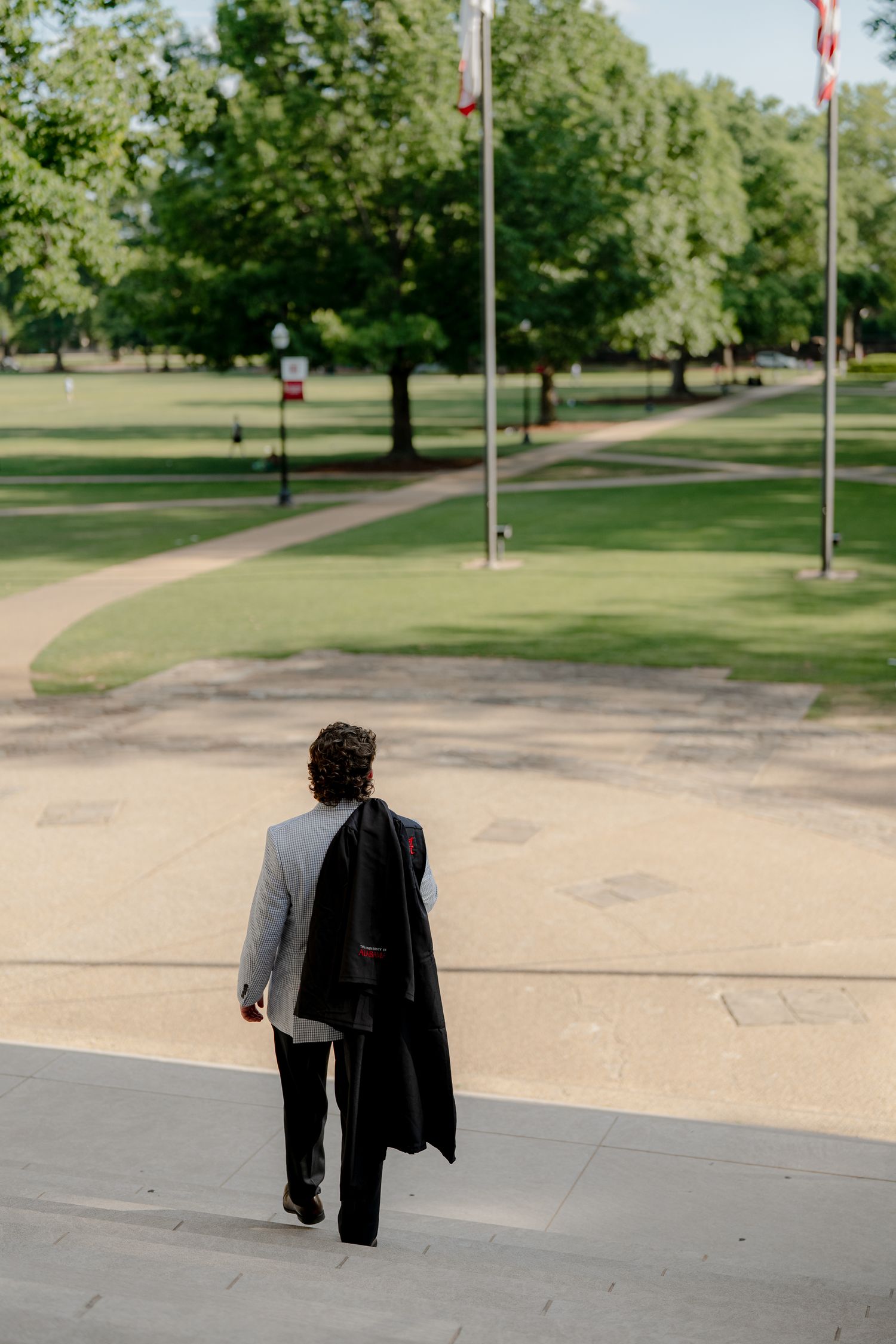 Man in gray blazer carrying black coat walks toward green park with flagpoles on sunny day.