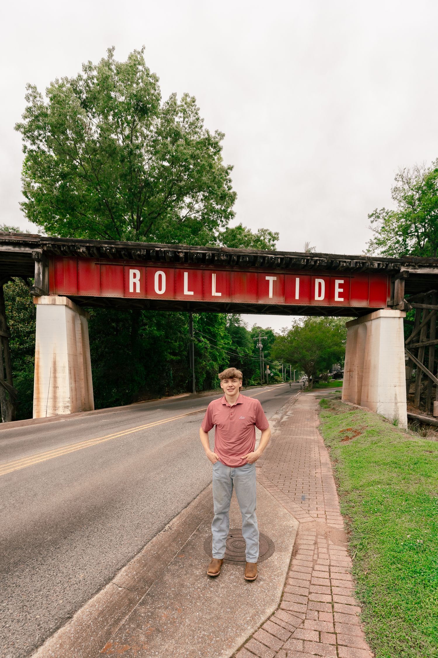 A person poses by a brick walkway under a red 'Roll Tide' sign on a bridge structure.