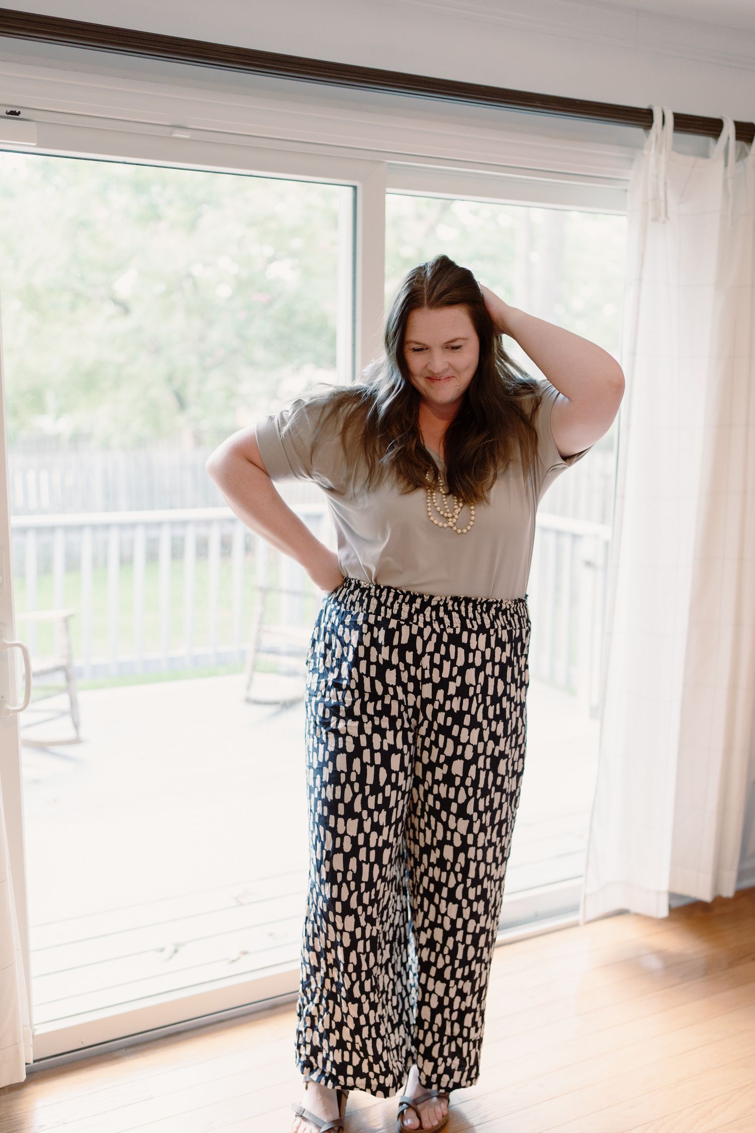 A person wearing a casual t-shirt and patterned skirt poses playfully in front of bright windows in a sunlit room.