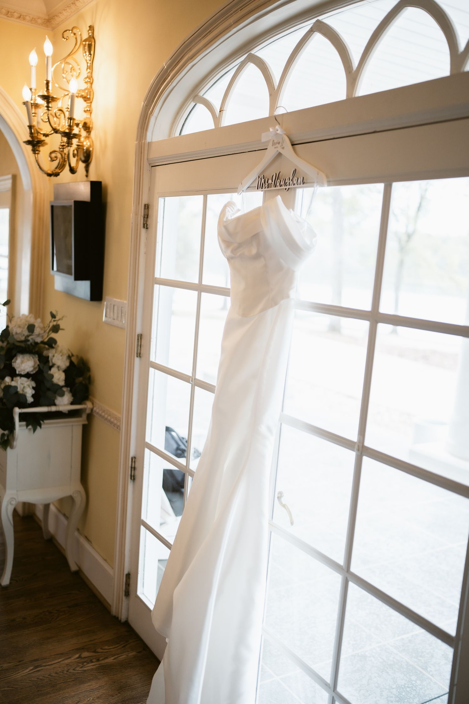 White wedding gown displayed on hanger against arched window with natural light streaming in.