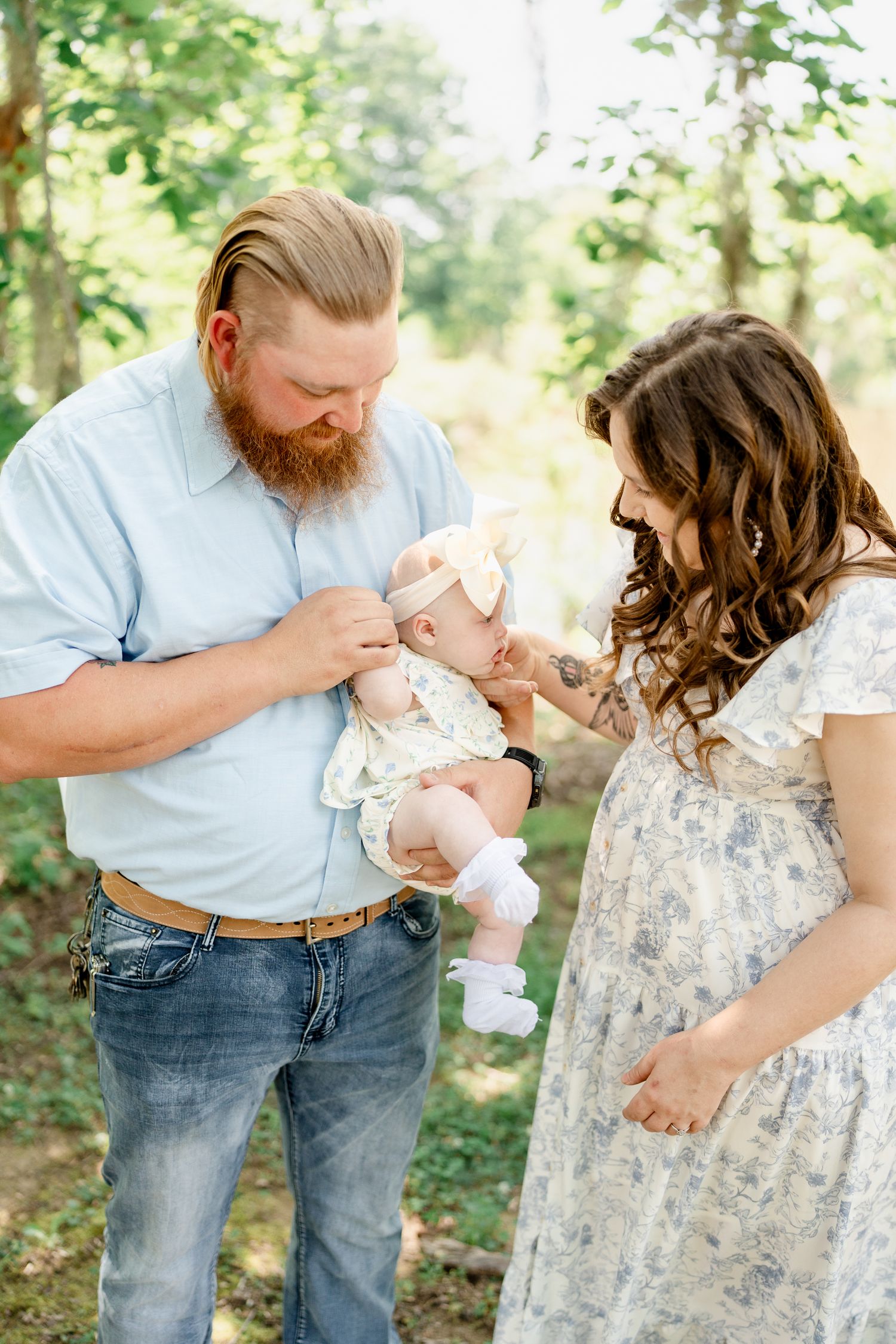 Family enjoying tender moments with baby in floral dress and light blue shirt in forest setting.