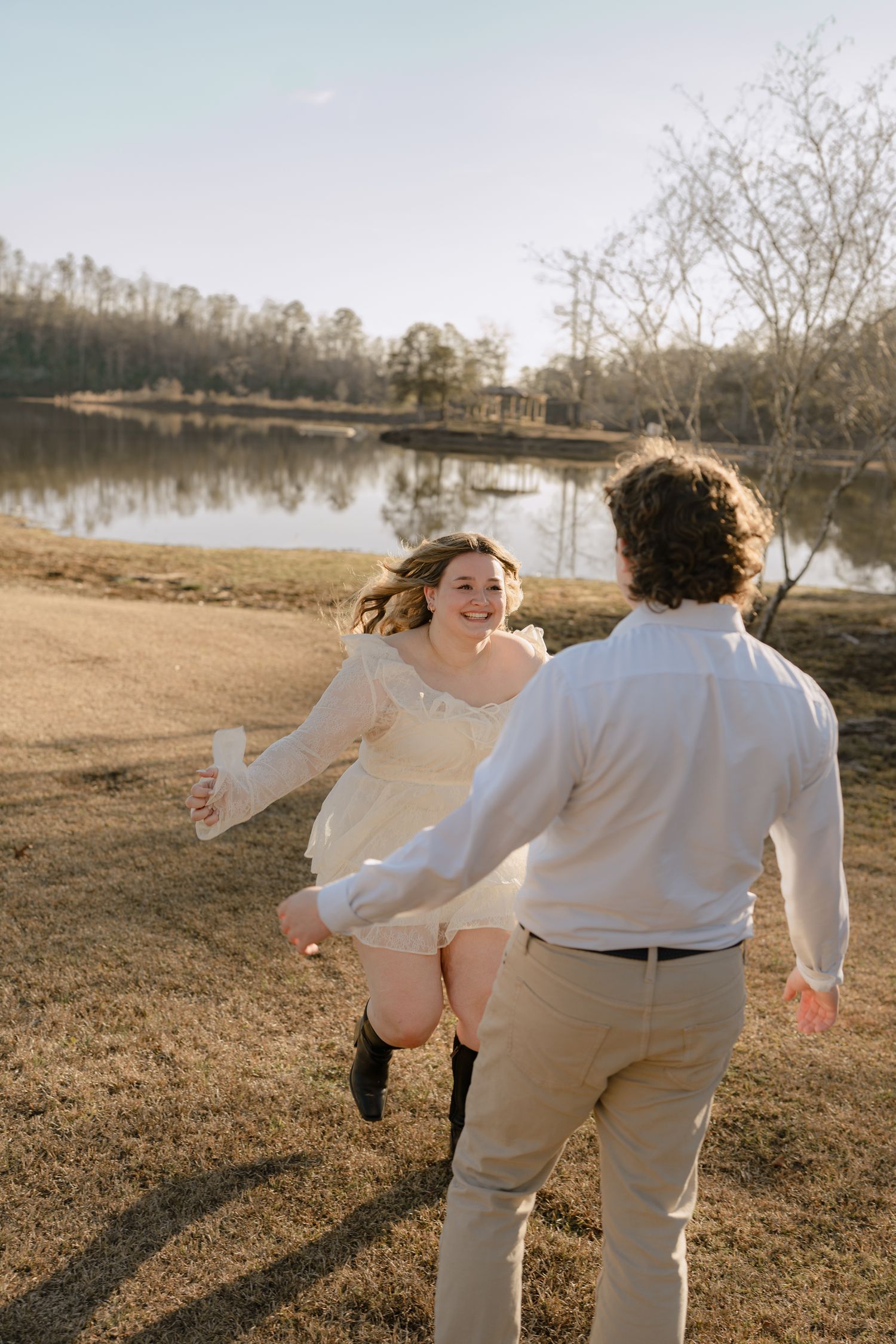 Joyful woman in white lace dress running toward her partner near a reflective lake.
