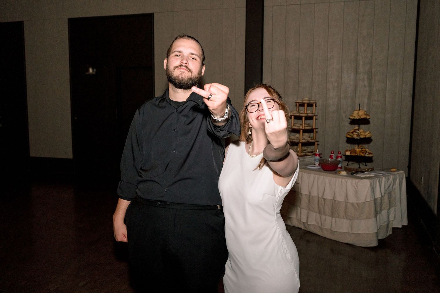 Couple at wedding reception showing off rings, man in black shirt and woman in white dress, cake table behind them.