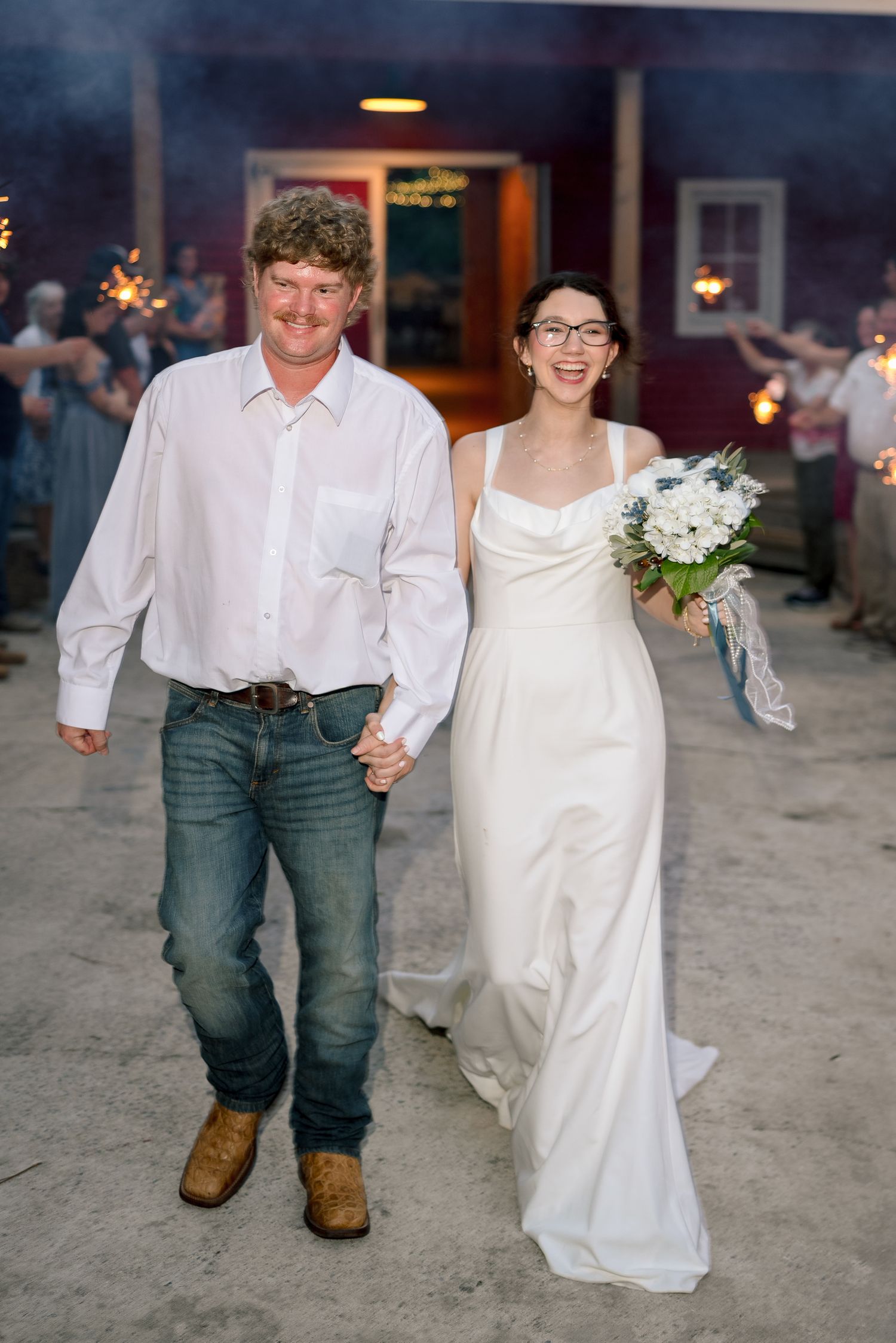 Bride and groom walk hand-in-hand through sparkler exit at rustic barn wedding, bride holding white floral bouquet.