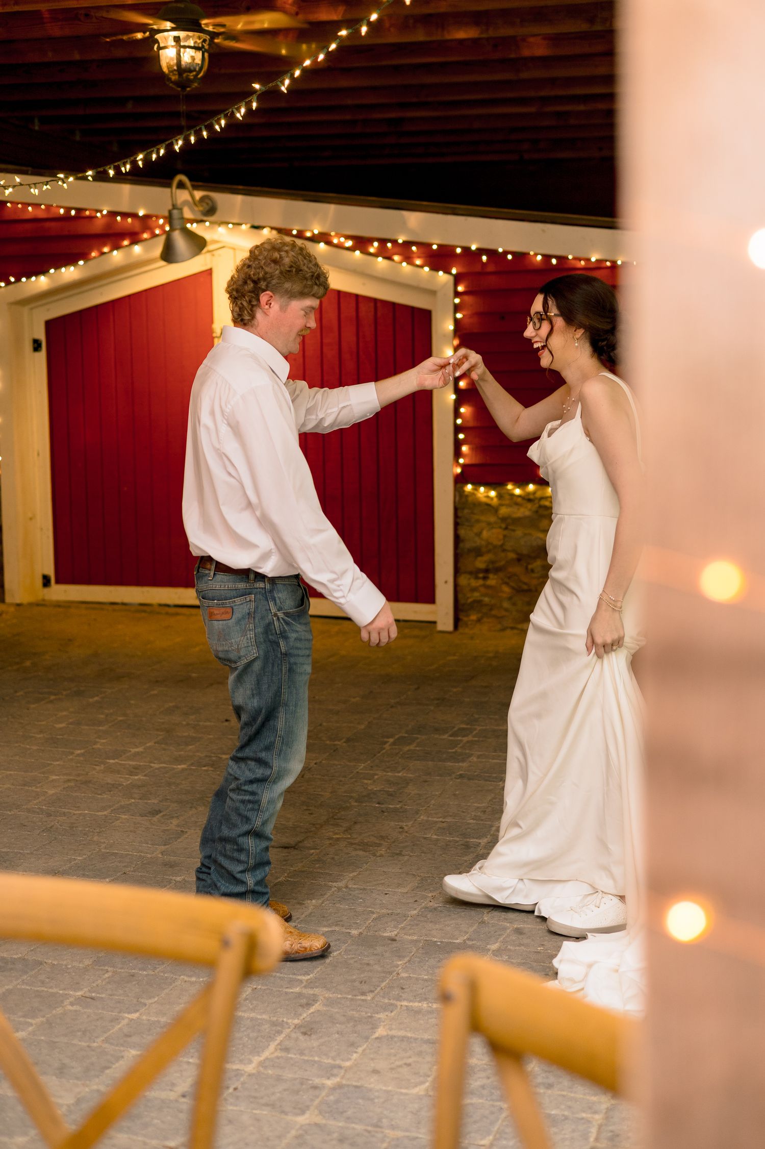 Bride and groom share a romantic first dance in a rustic barn venue decorated with warm string lights.