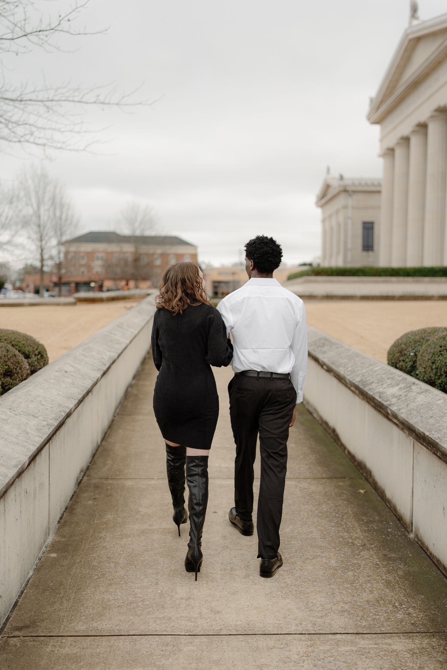 Couple walking arm in arm down a formal pathway toward a neoclassical building on a cloudy day.
