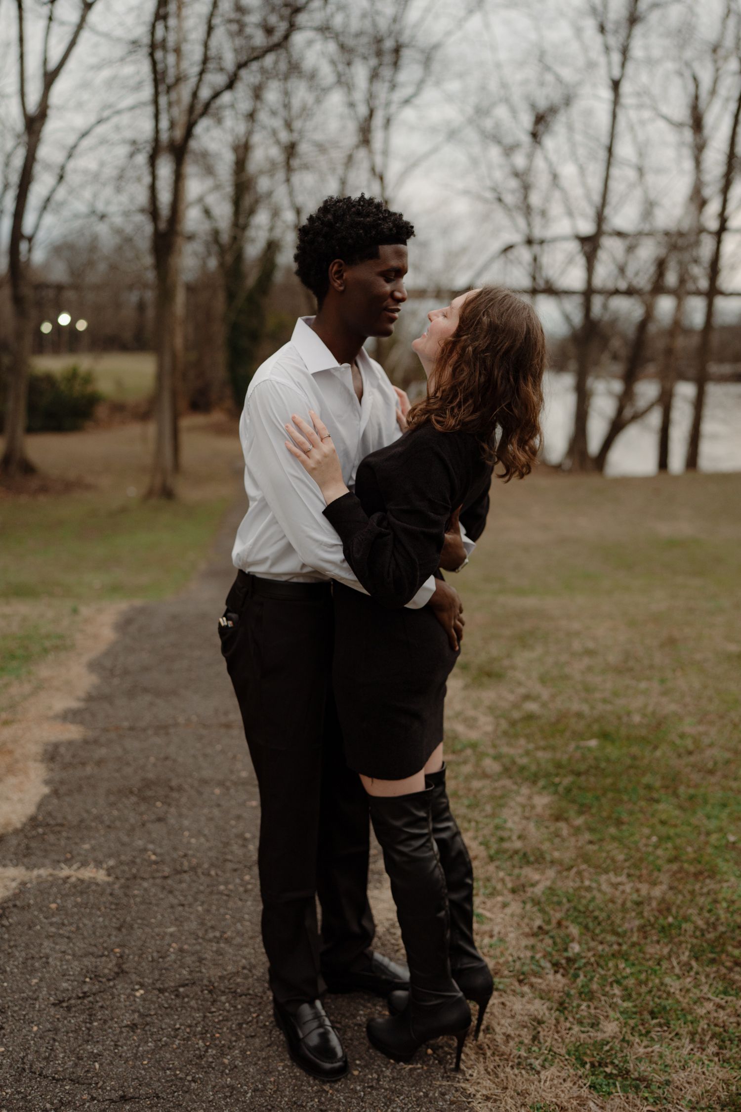 An interracial couple embraces and gazes at each other on a gravel path surrounded by bare winter trees.