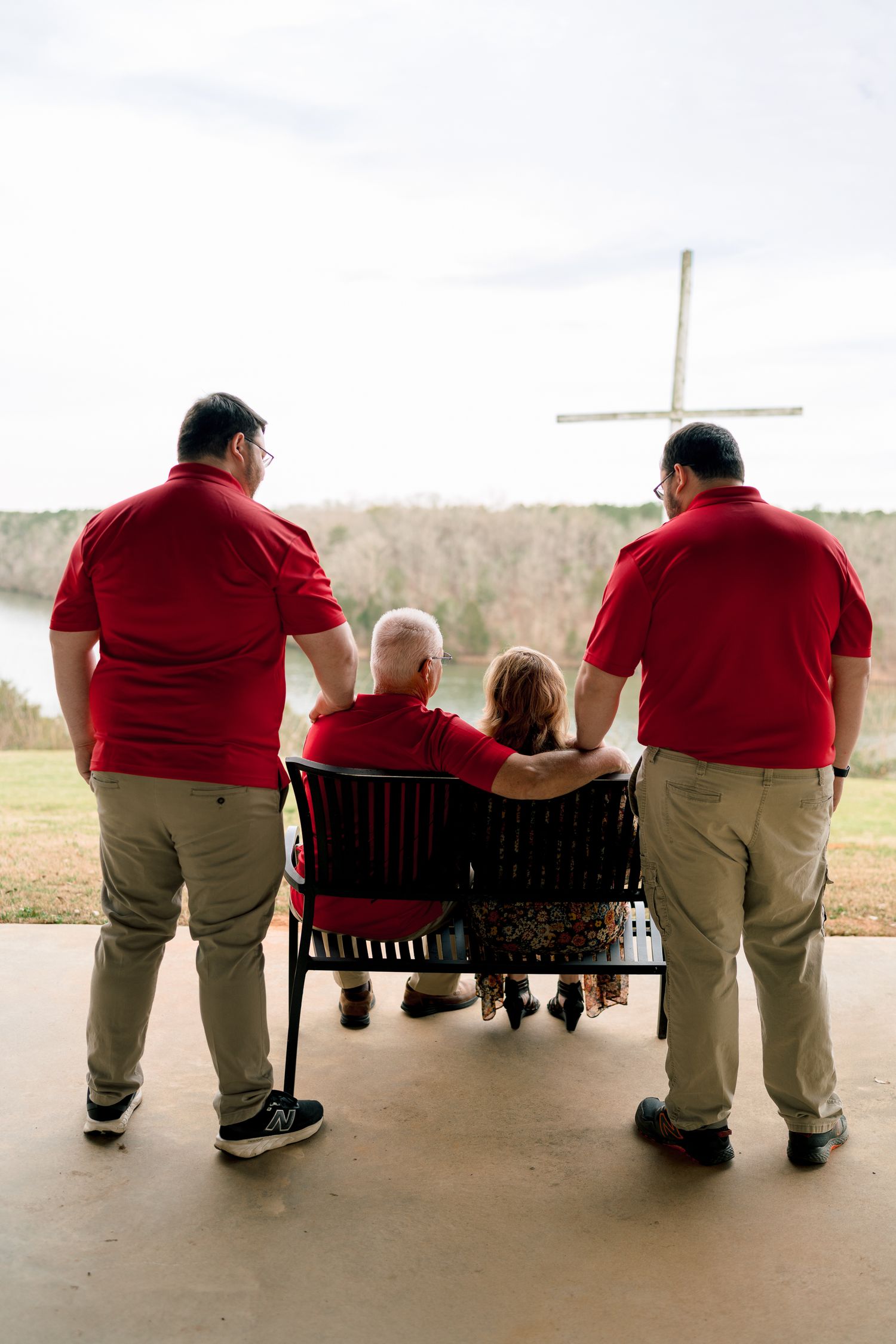 Family in matching red shirts gather around elderly couple on bench overlooking a cross and lake.