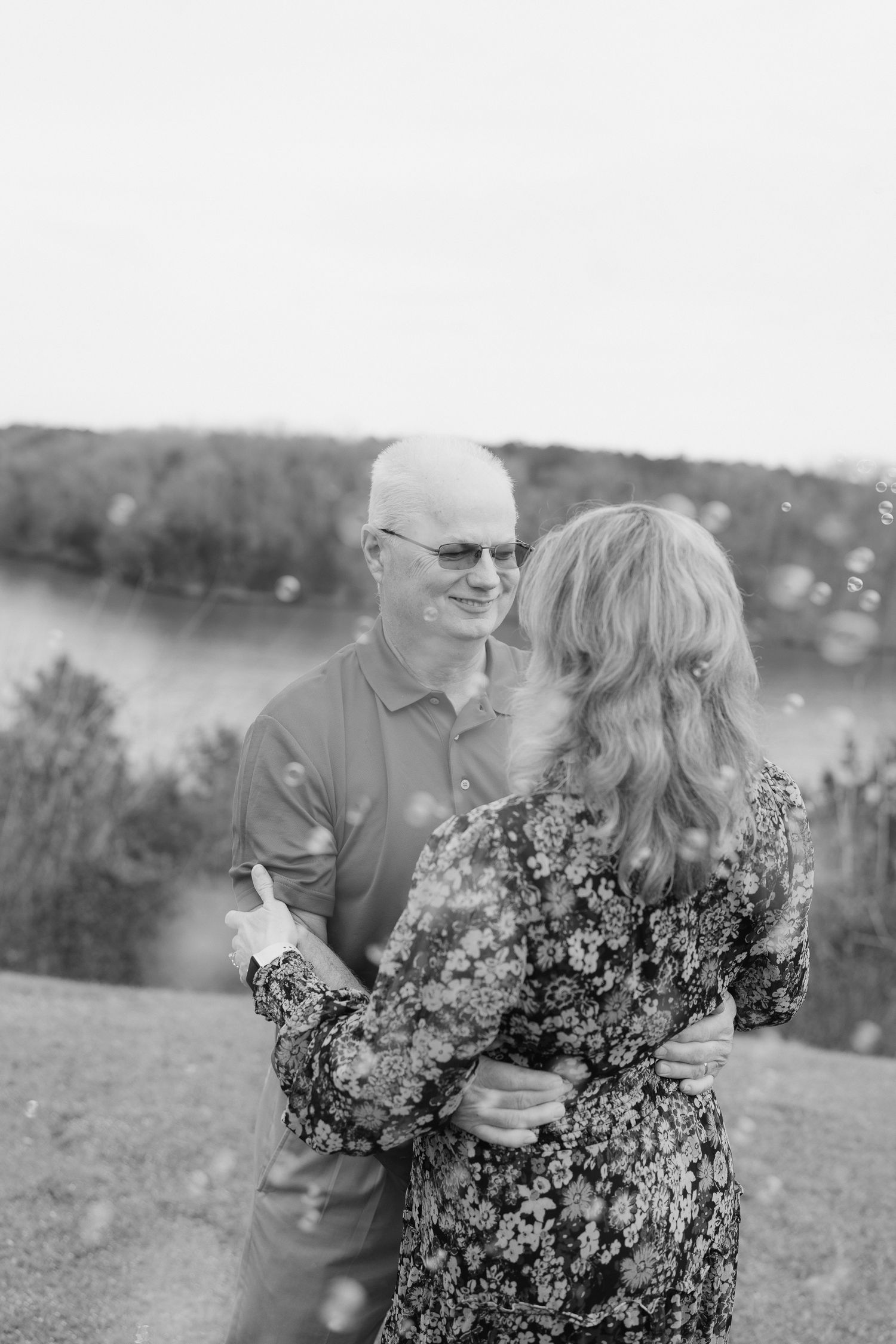 Black and white photo of older couple embracing and smiling near a misty lake.