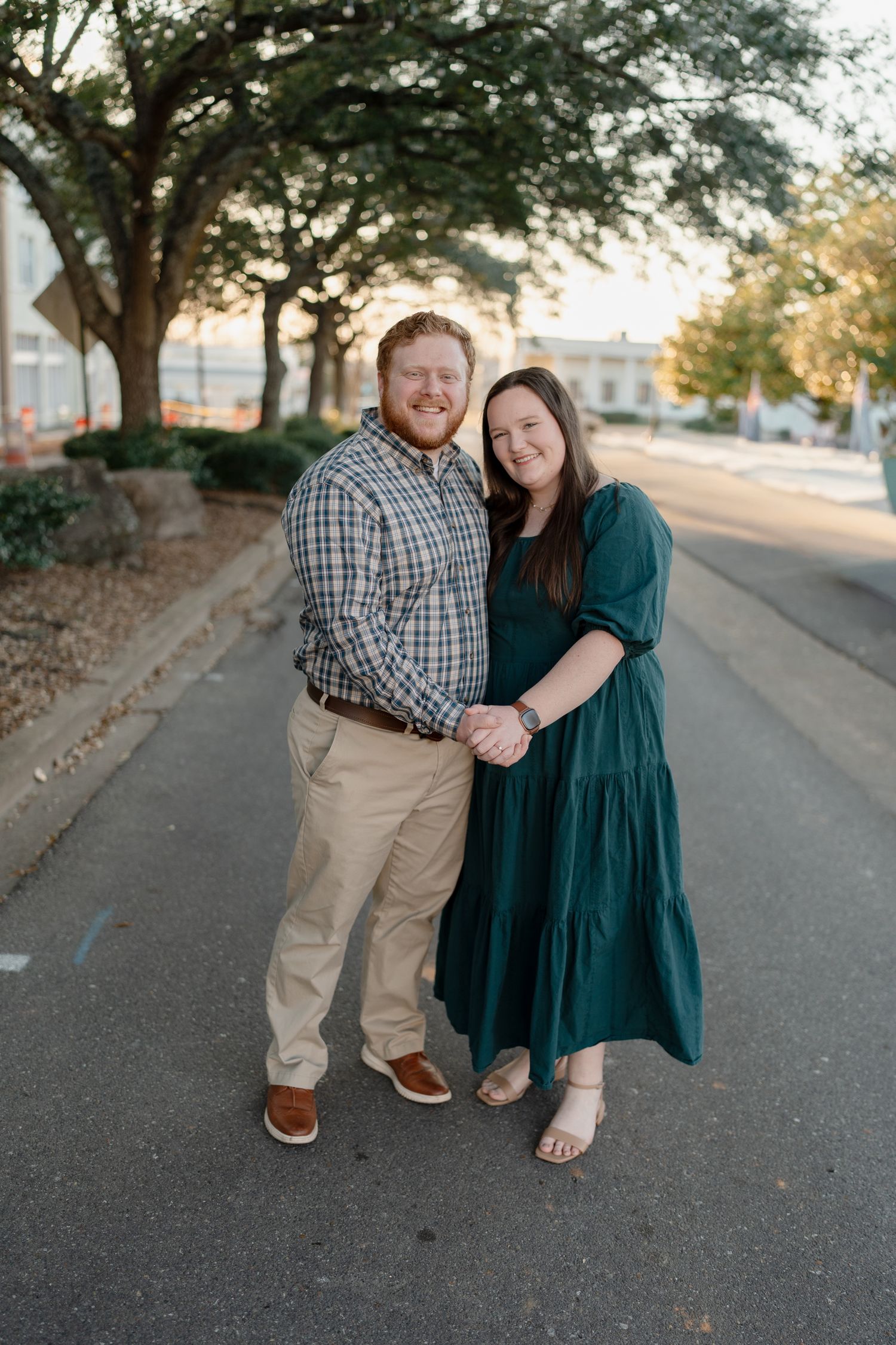 Couple holding hands and smiling on a tree-lined street during golden hour engagement session.