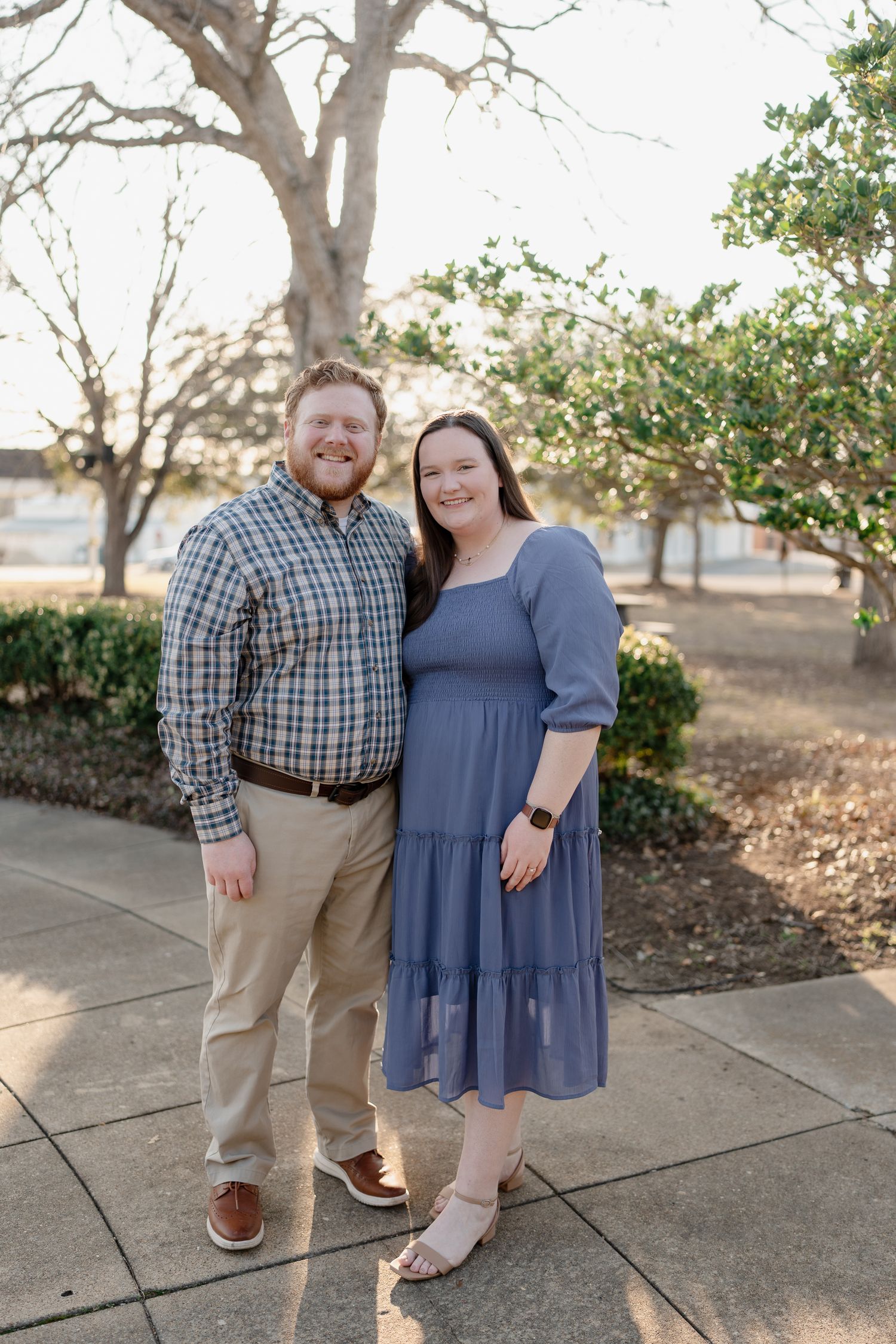 A smiling couple poses outdoors on a sidewalk, man in plaid shirt and khakis, woman in blue tiered dress.