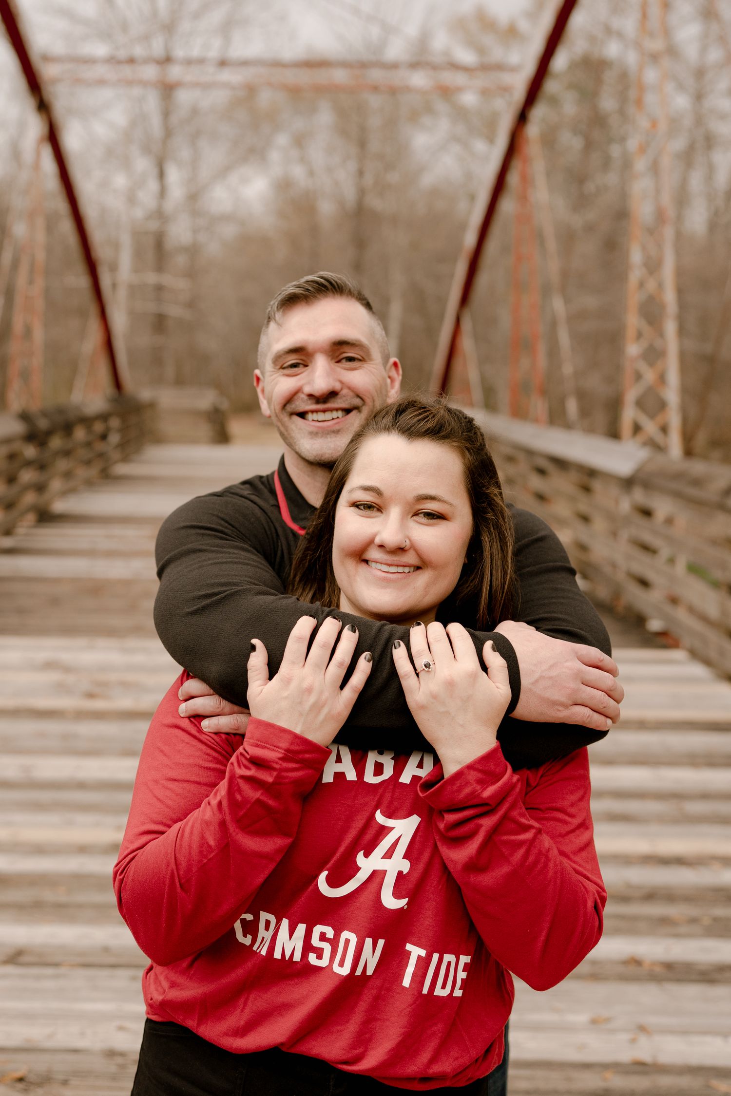 A couple in Alabama Crimson Tide attire embracing on a wooden bridge during an autumn day.