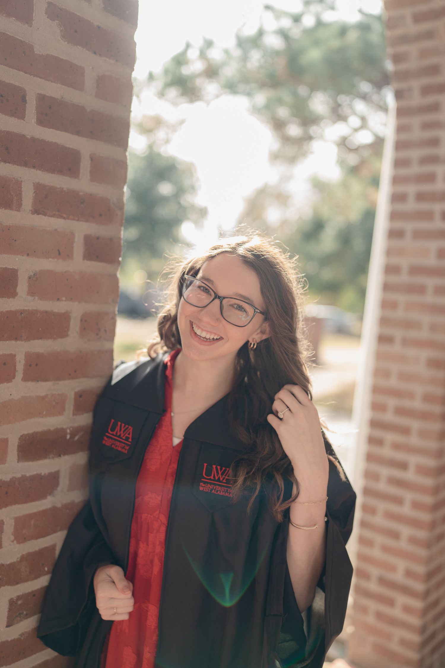 Student in graduation attire laughs joyfully while posing between brick columns in natural sunlight.