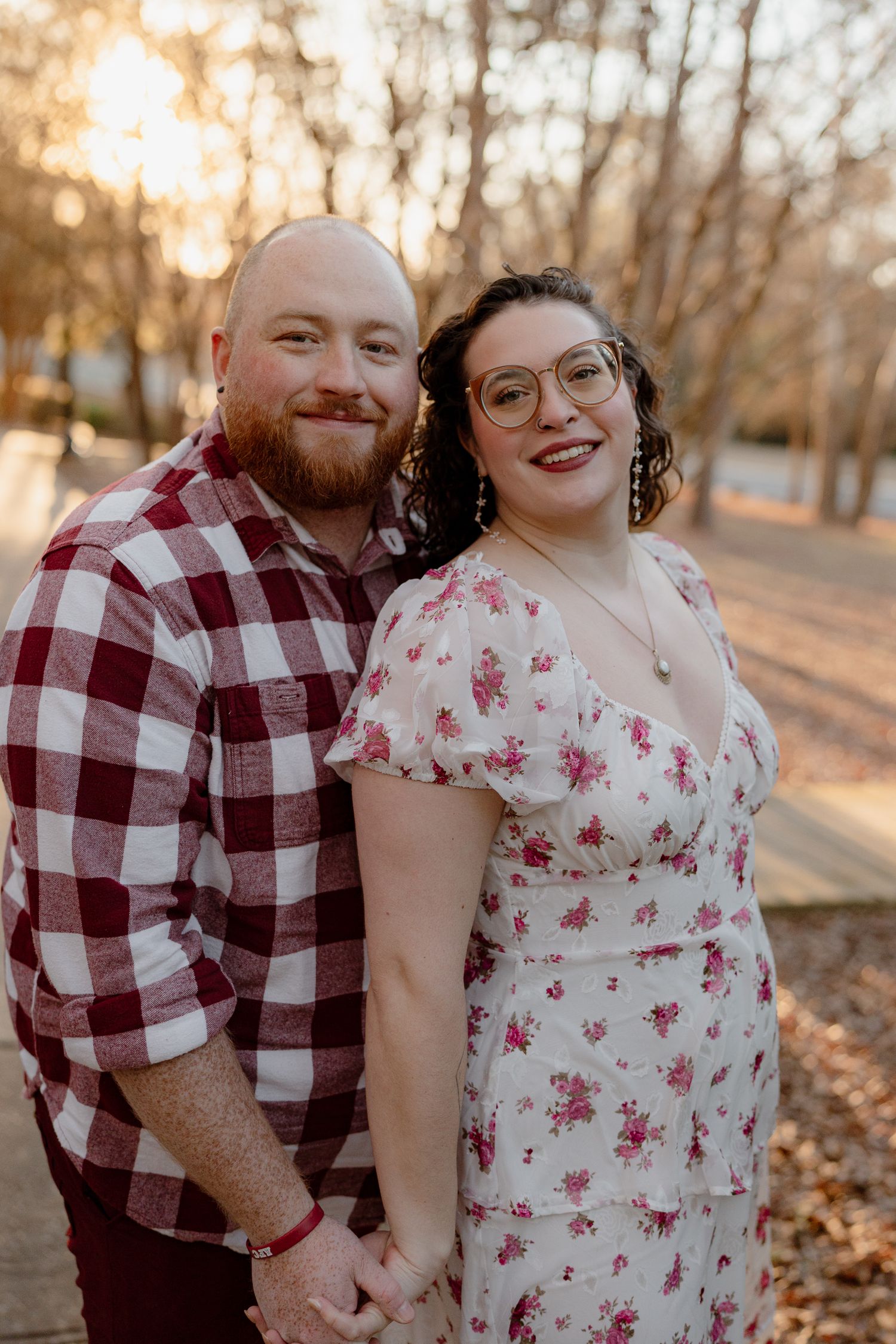 A couple embraces and laughs together while standing in a park with fallen leaves during golden hour.