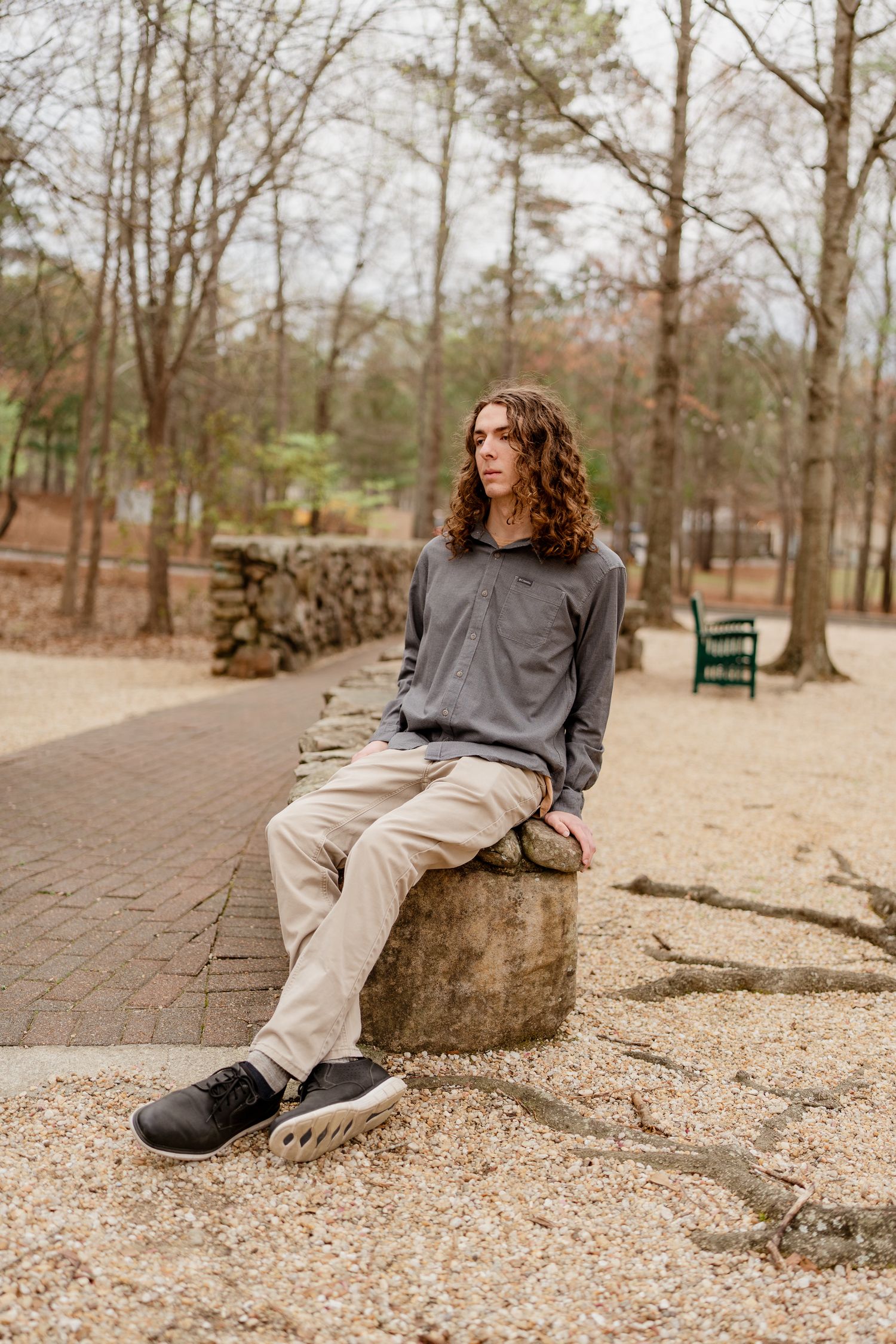 Person in casual attire sitting on stone wall along brick pathway in wooded park setting.