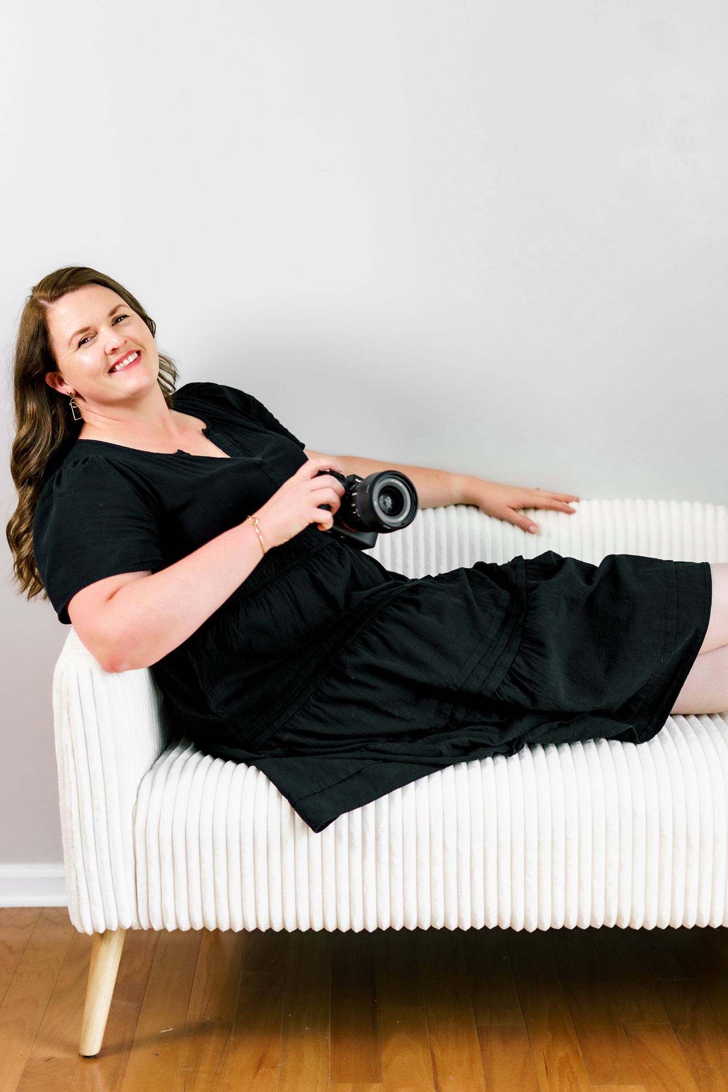 Person in black dress lounges playfully on white bench while holding camera.